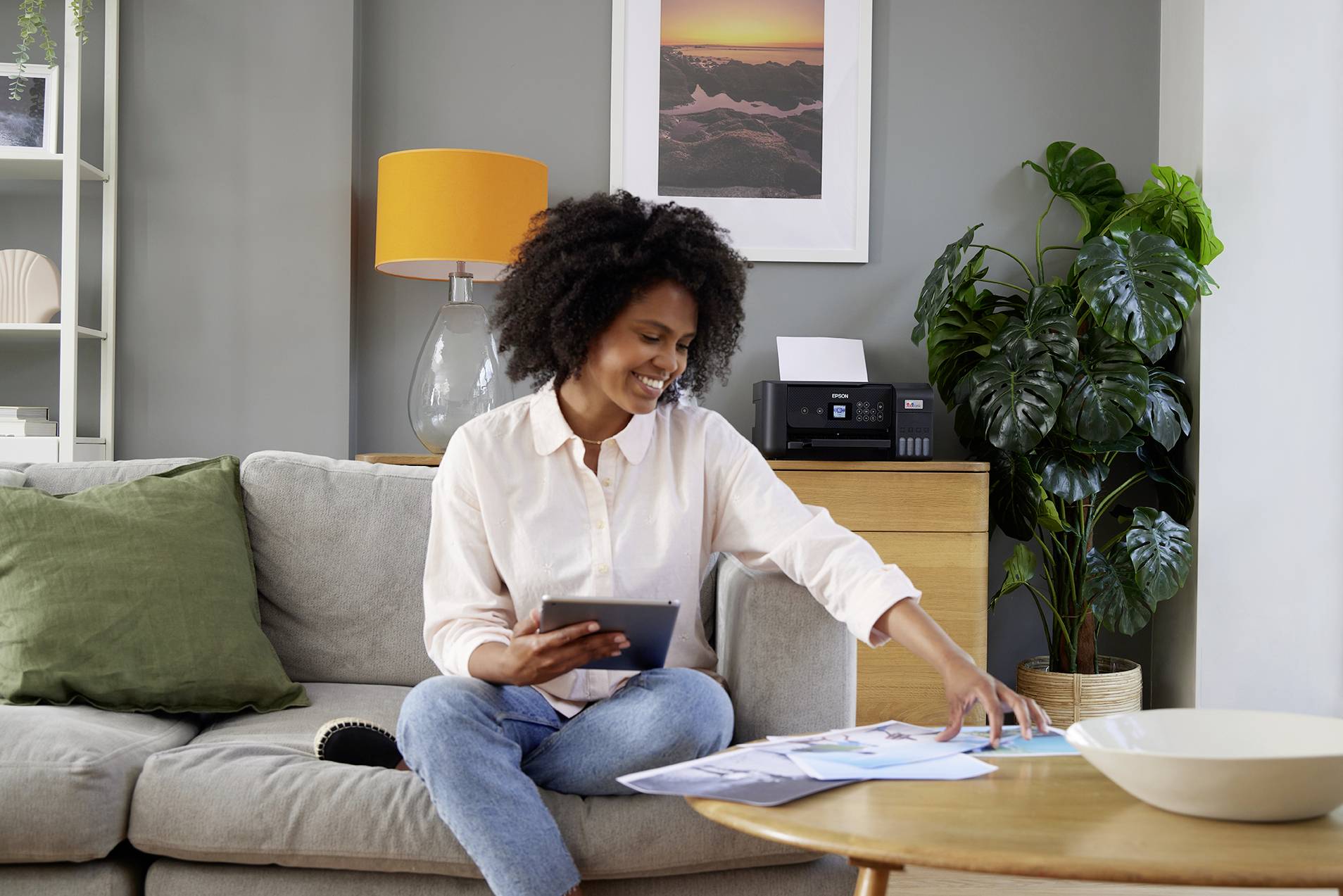 A woman is sitting on a sofa, holding a tablet and examining documents on a table. Living room with a potted plant and lamp in the background.