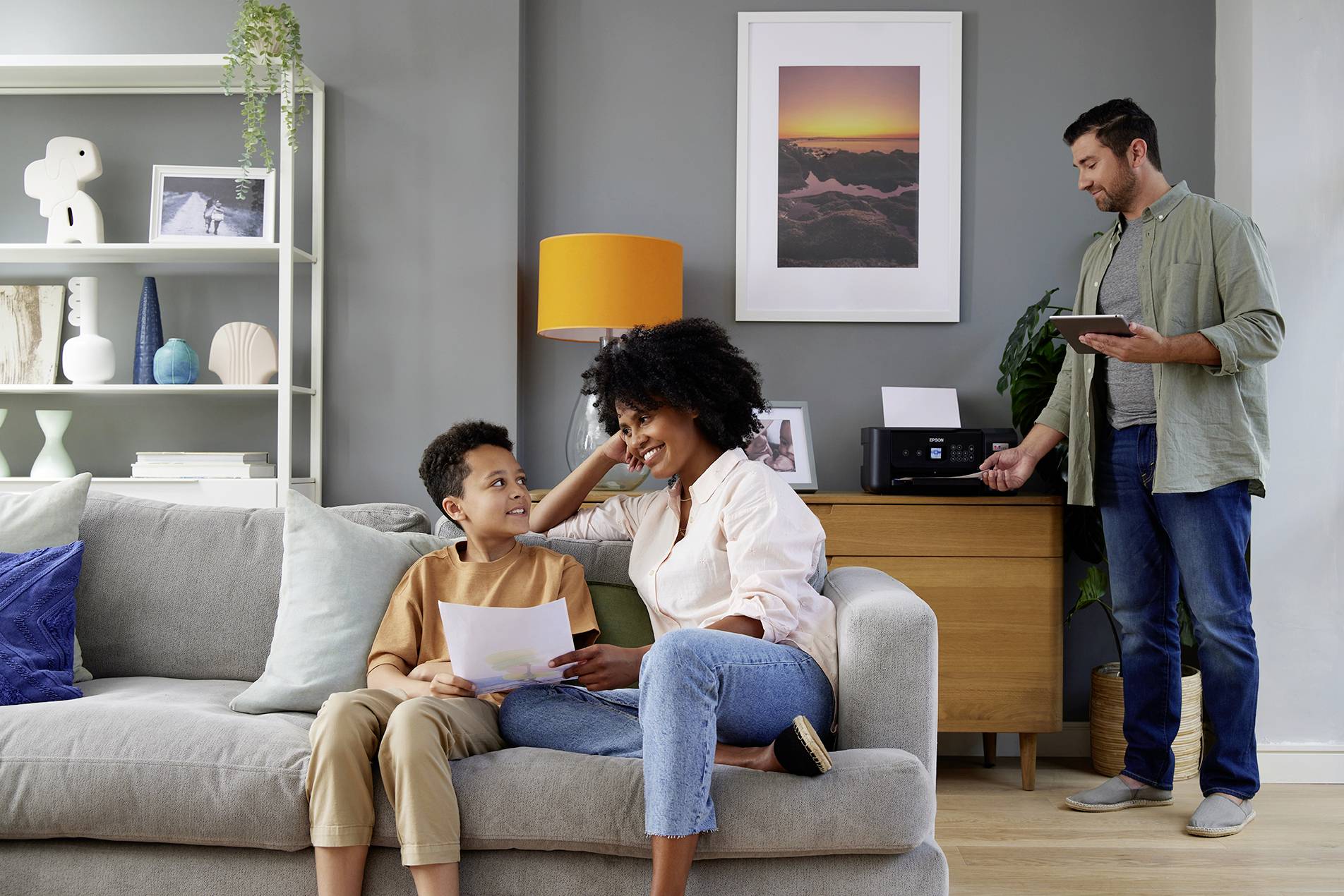 A woman and a boy are sitting smiling on a sofa, looking at a photograph. A man is standing beside them, holding a tablet. A living room is in the background.