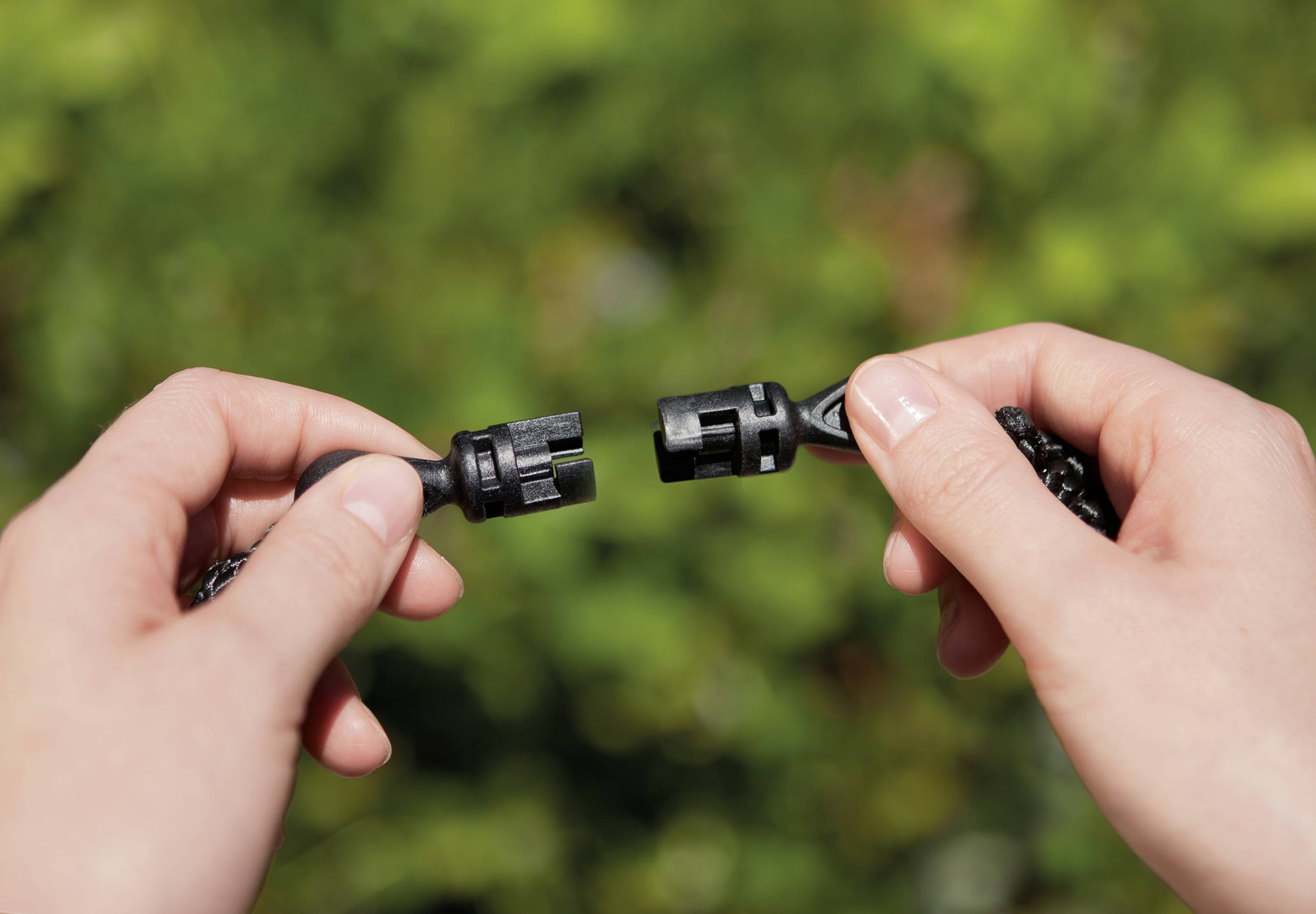 One hand holds a male connector on the left, the other hand holds a female connector on the right. Both connectors are about to be connected. Green leaves can be seen blurred in the background.