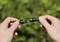 One hand holds a male connector on the left, the other hand holds a female connector on the right. Both connectors are about to be connected. Green leaves can be seen blurred in the background.