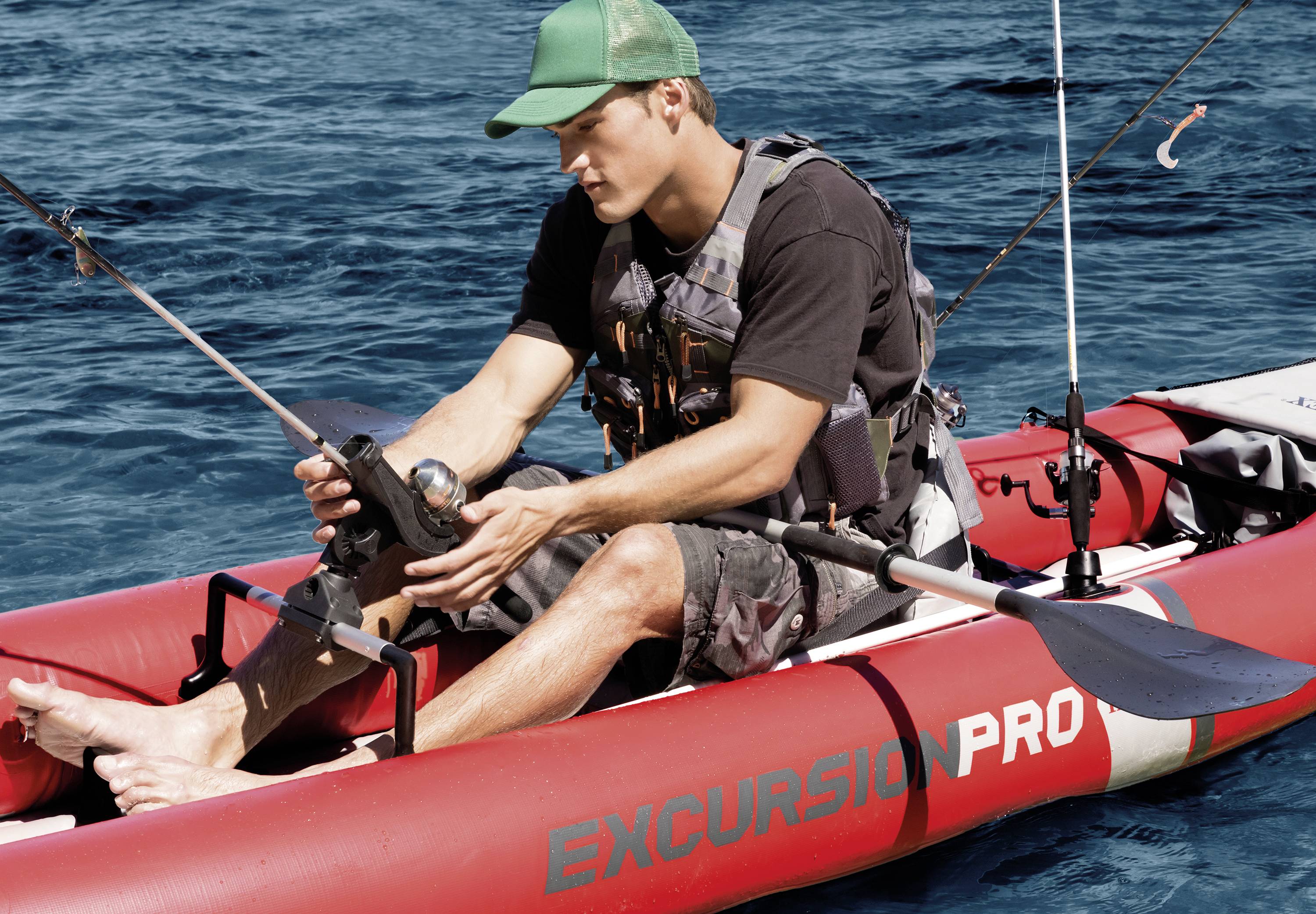 A man is fishing on a red inflatable dinghy with 'EXCURSION PRO' written on it, floating on blue water. He is wearing a green cap and a life jacket.