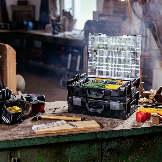 A cluttered workbench in a workshop with tools, a toolbox, and various small containers scattered around, suggesting active work in progress.