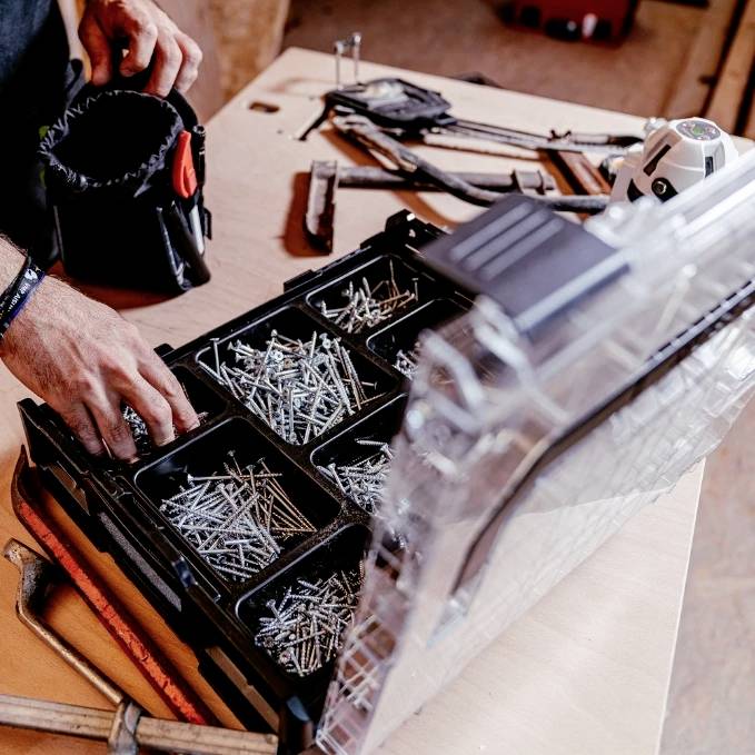 A person organizing screws in a toolbox on a workbench, with various tools and a measuring tape nearby.