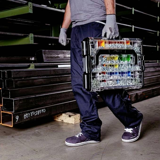 A person wearing gloves holds a plastic toolkit with colored compartments, standing next to metal shelving.