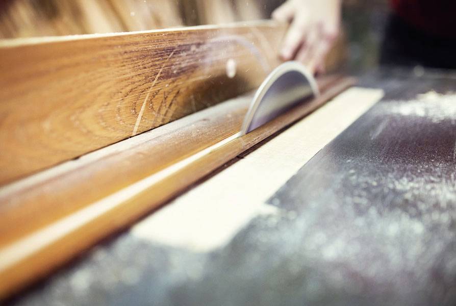 A hand guides a wooden board against a rotating saw blade on a table to cut it. Sawdust lies on the surface.