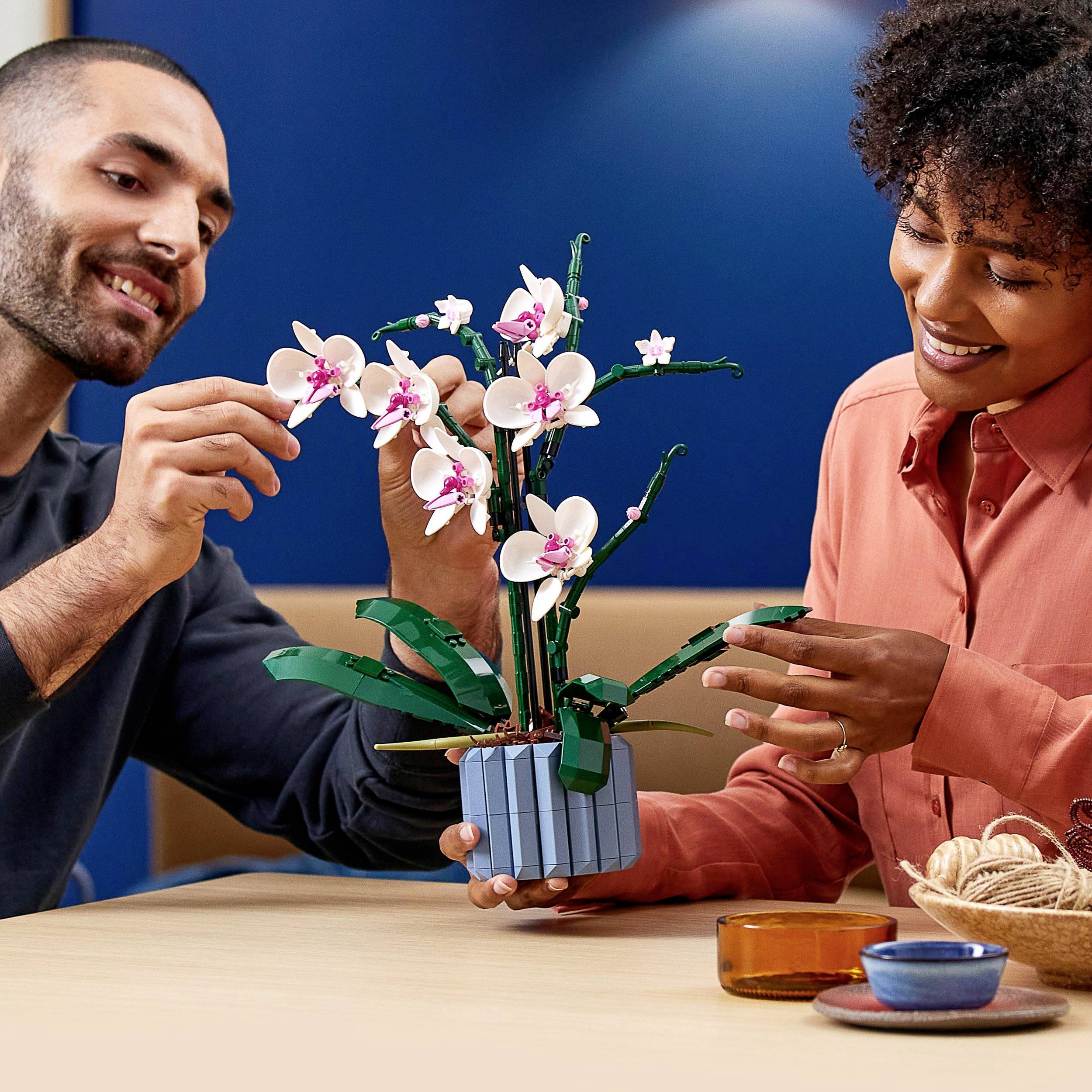 A man and a woman are sitting at a table and smiling while building an orchid plant together using LEGO bricks.