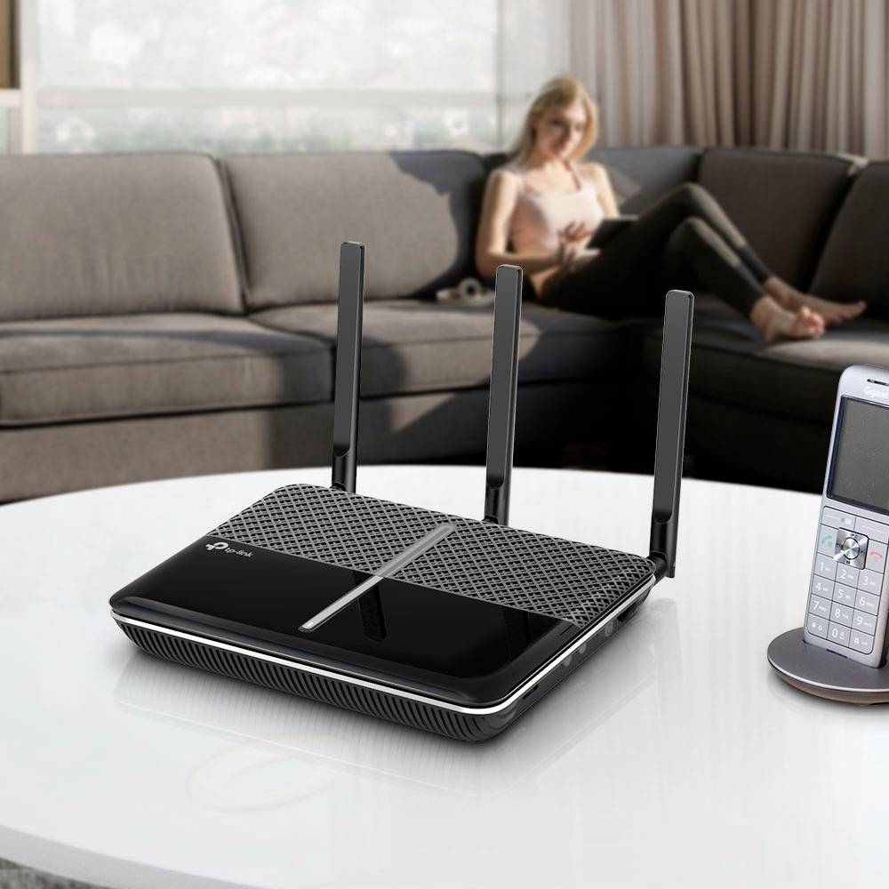 A Wi-Fi router with three aerials sits on a table in the living room; in the background, a person is seated on a sofa.