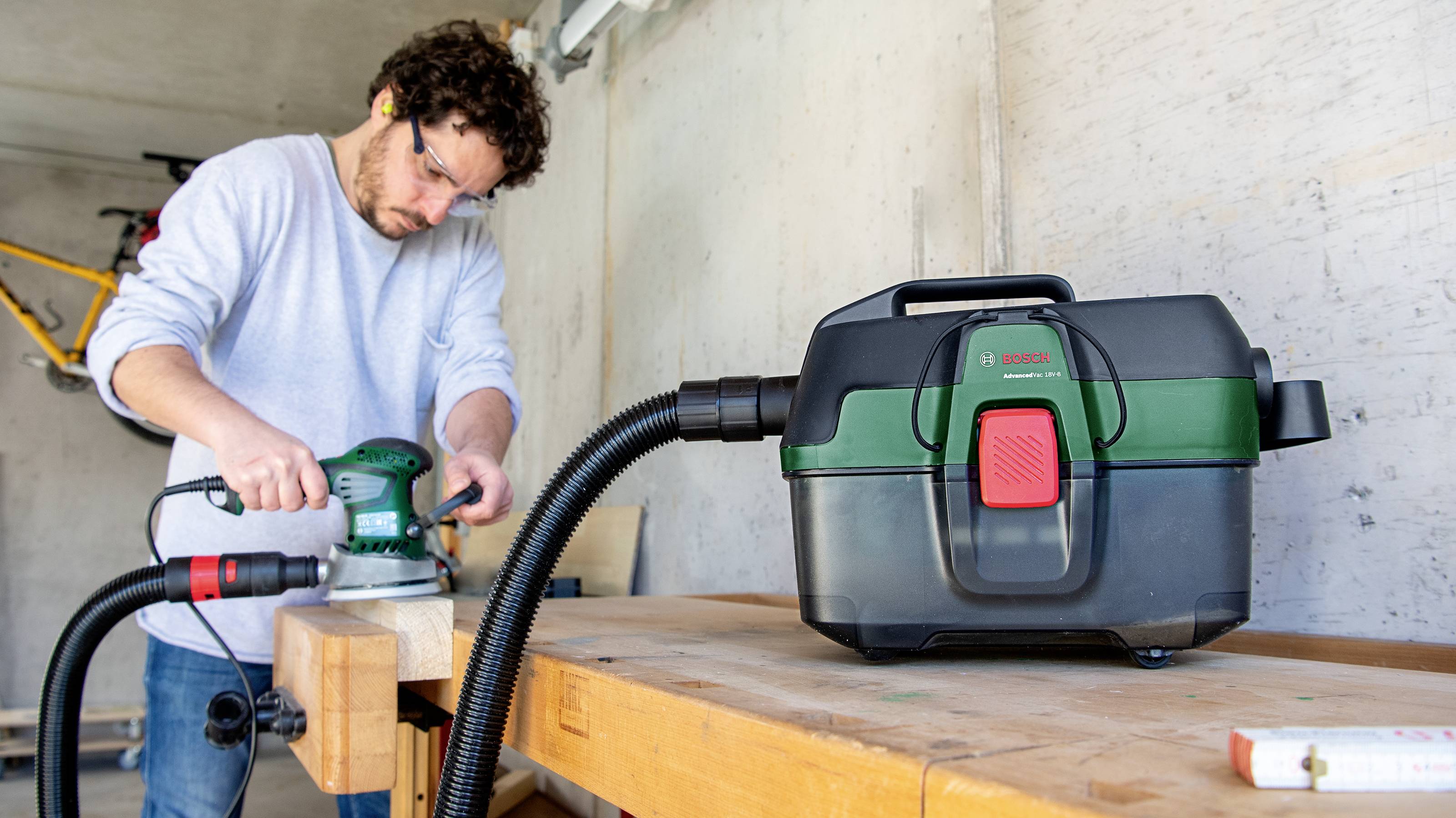 A man is sanding wood on a workbench, with a green and black industrial vacuum cleaner standing nearby.