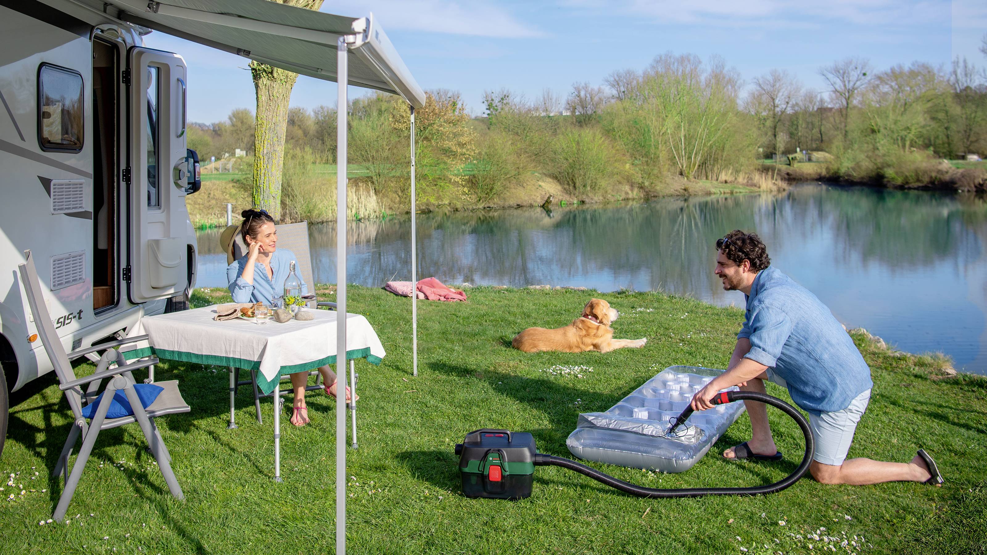 A man is inflating an air bed next to a camper van, while a woman sits at a table. In the background, a dog is lying down and a pond can be seen.