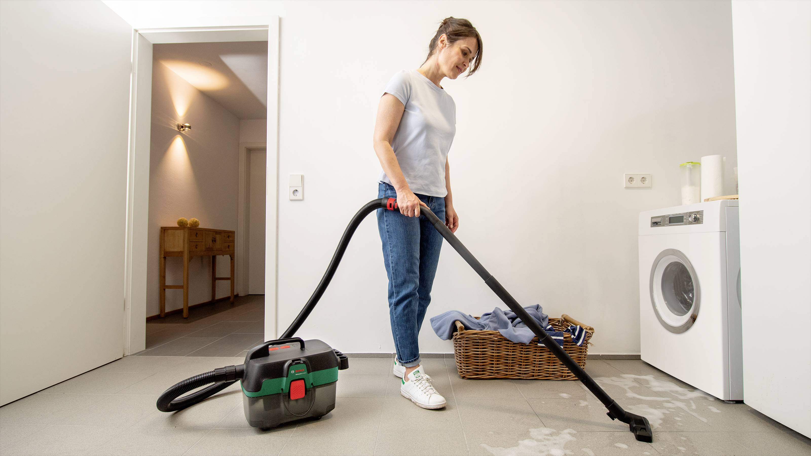 A woman is cleaning up water with a wet and dry vacuum cleaner on a floor near a washing machine.