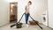 A woman is cleaning up water with a wet and dry vacuum cleaner on a floor near a washing machine.