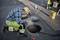 A worker in safety clothing inspects the opening of a manhole cover on a street, surrounded by traffic cones, using a torch.