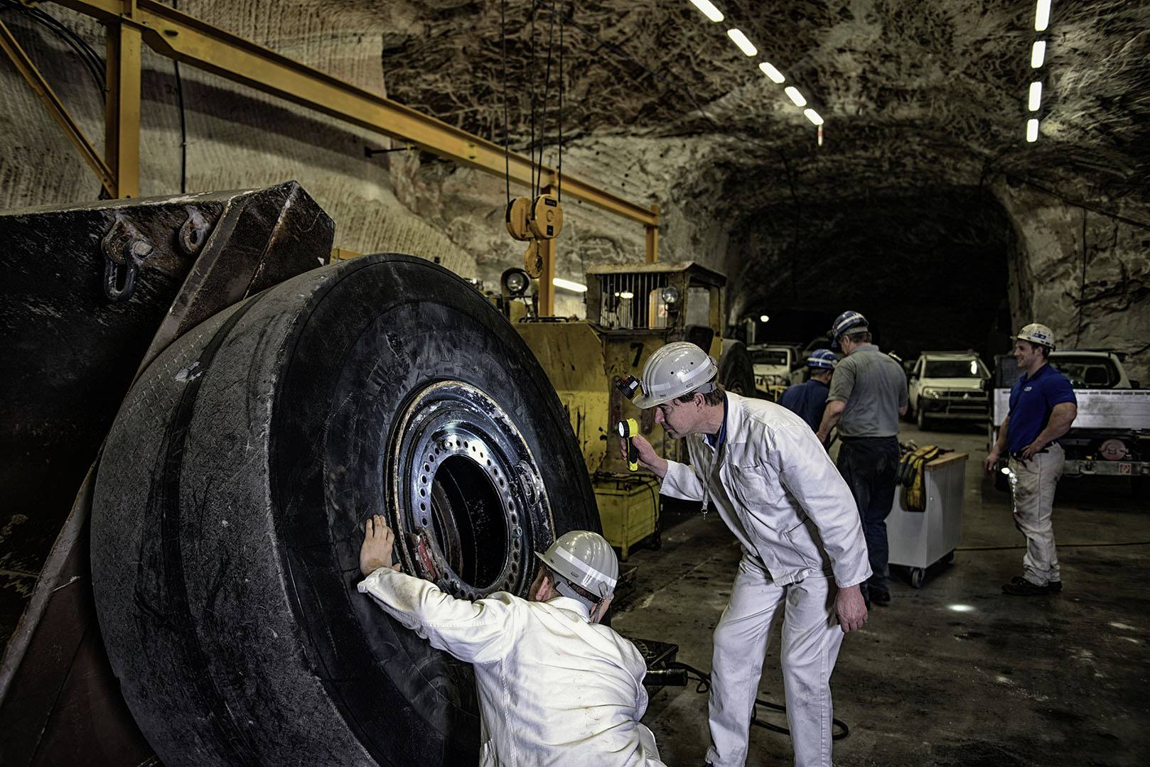 Workers in a mine are repairing a massive machine wheel. In the background, a dark tunnel can be seen.