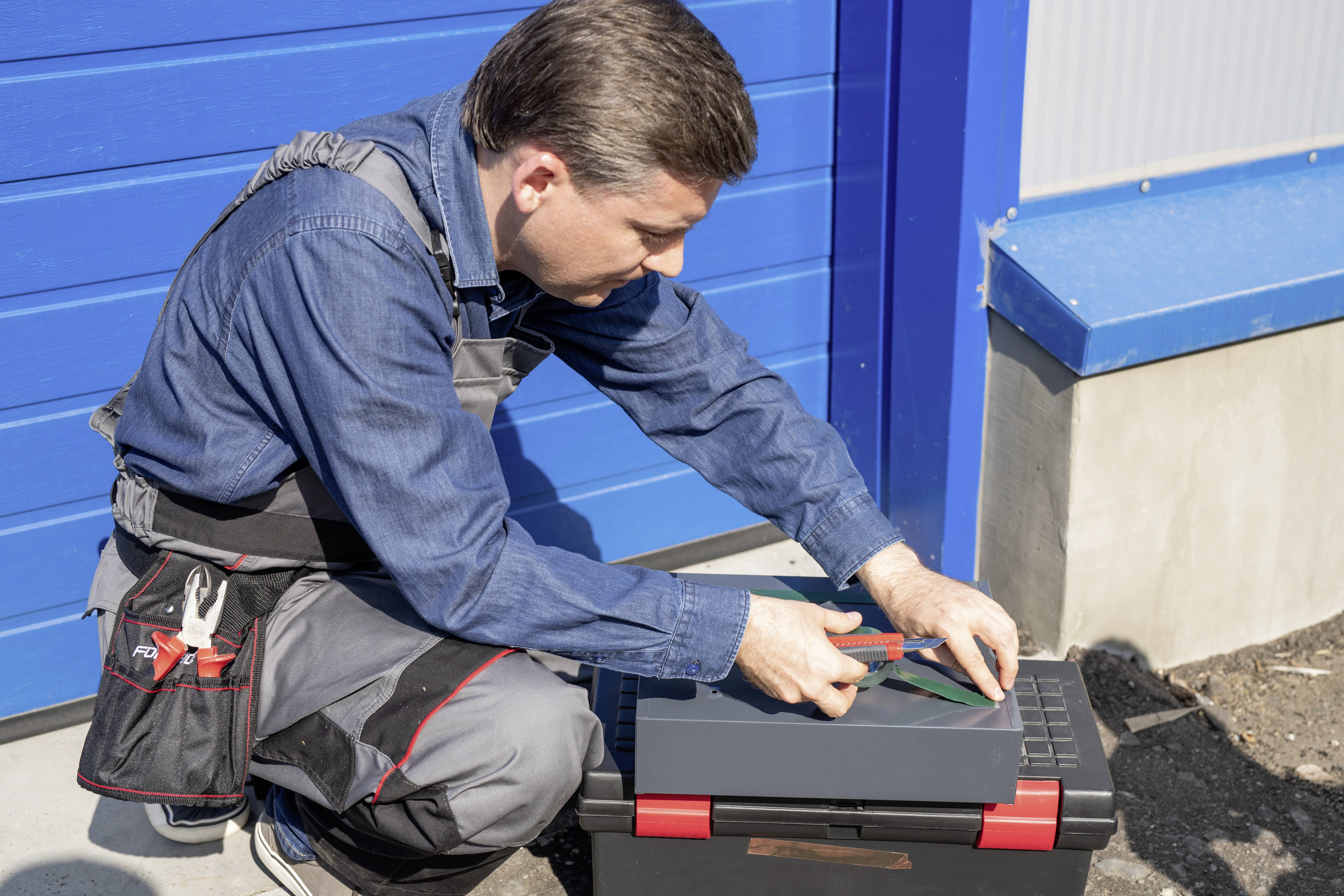 A man in workwear is sitting on a toolbox in front of a blue garage door, cutting something with a Stanley knife.