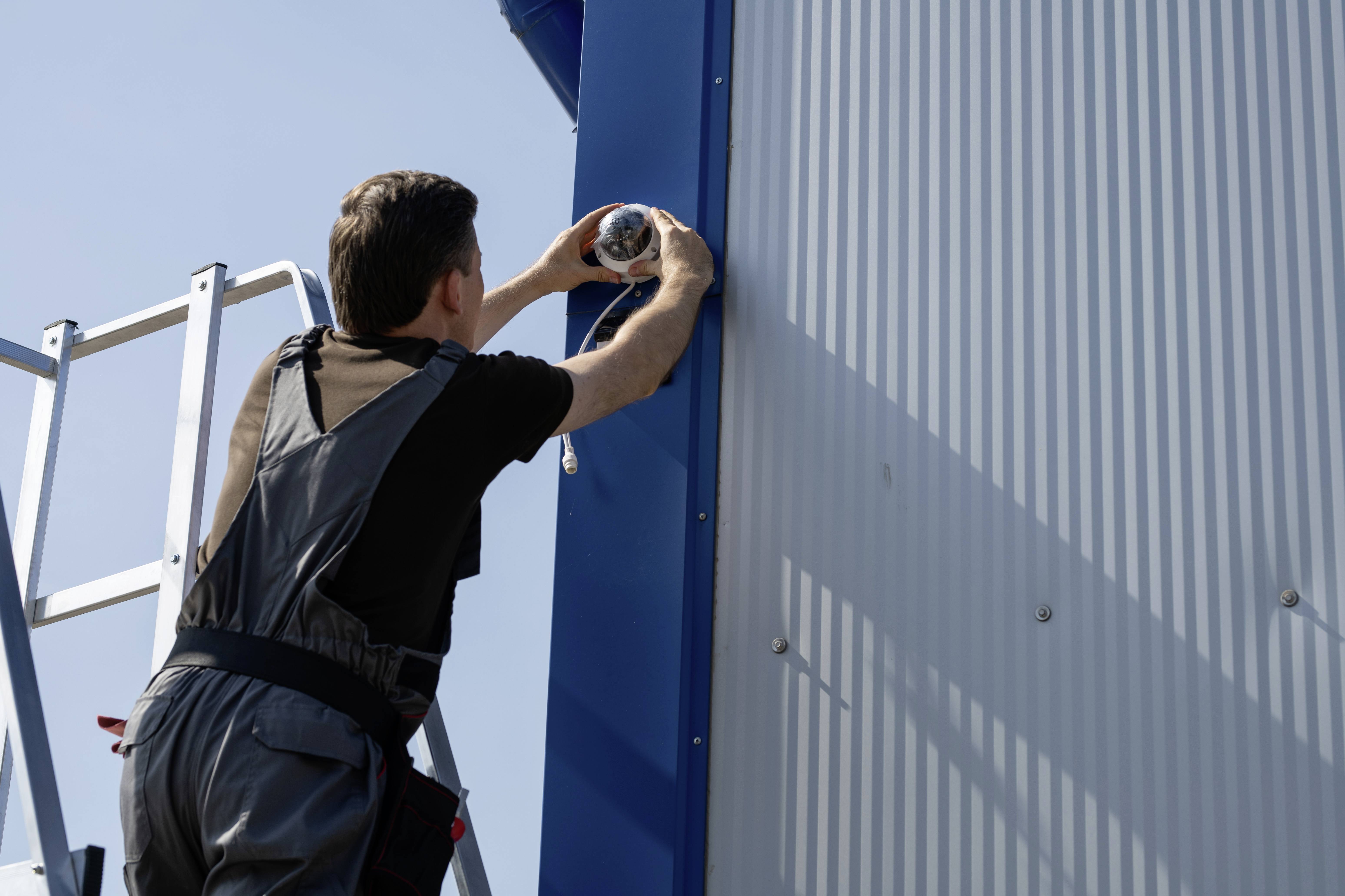 A man is installing a surveillance camera on the external side wall of a building, standing on a ladder under a clear sky.
