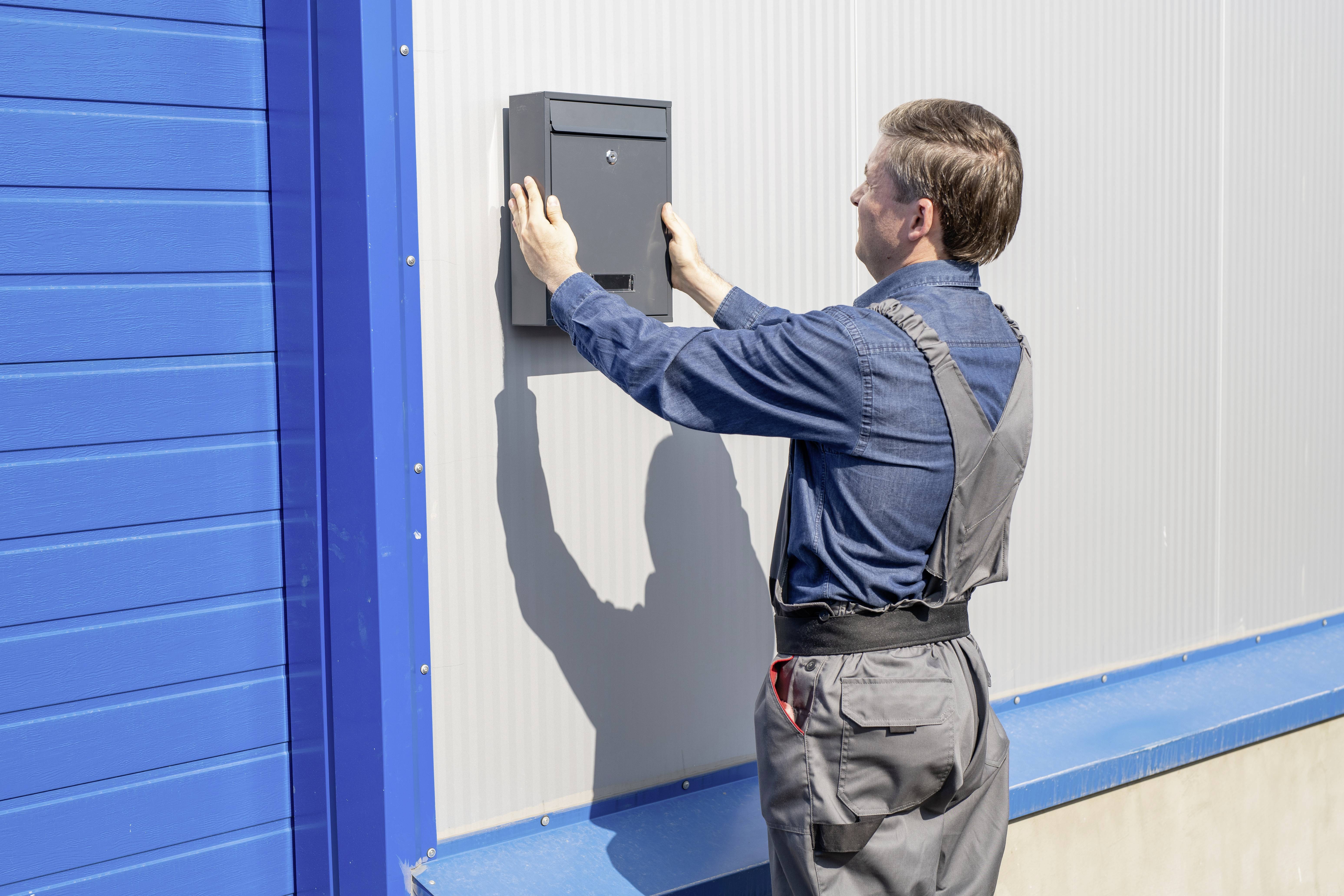 A man is installing a postbox on a metal exterior wall. He is wearing work attire and is focused on his task.