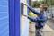 A man in workwear is installing an electronic box on the wall of a blue building outdoors.