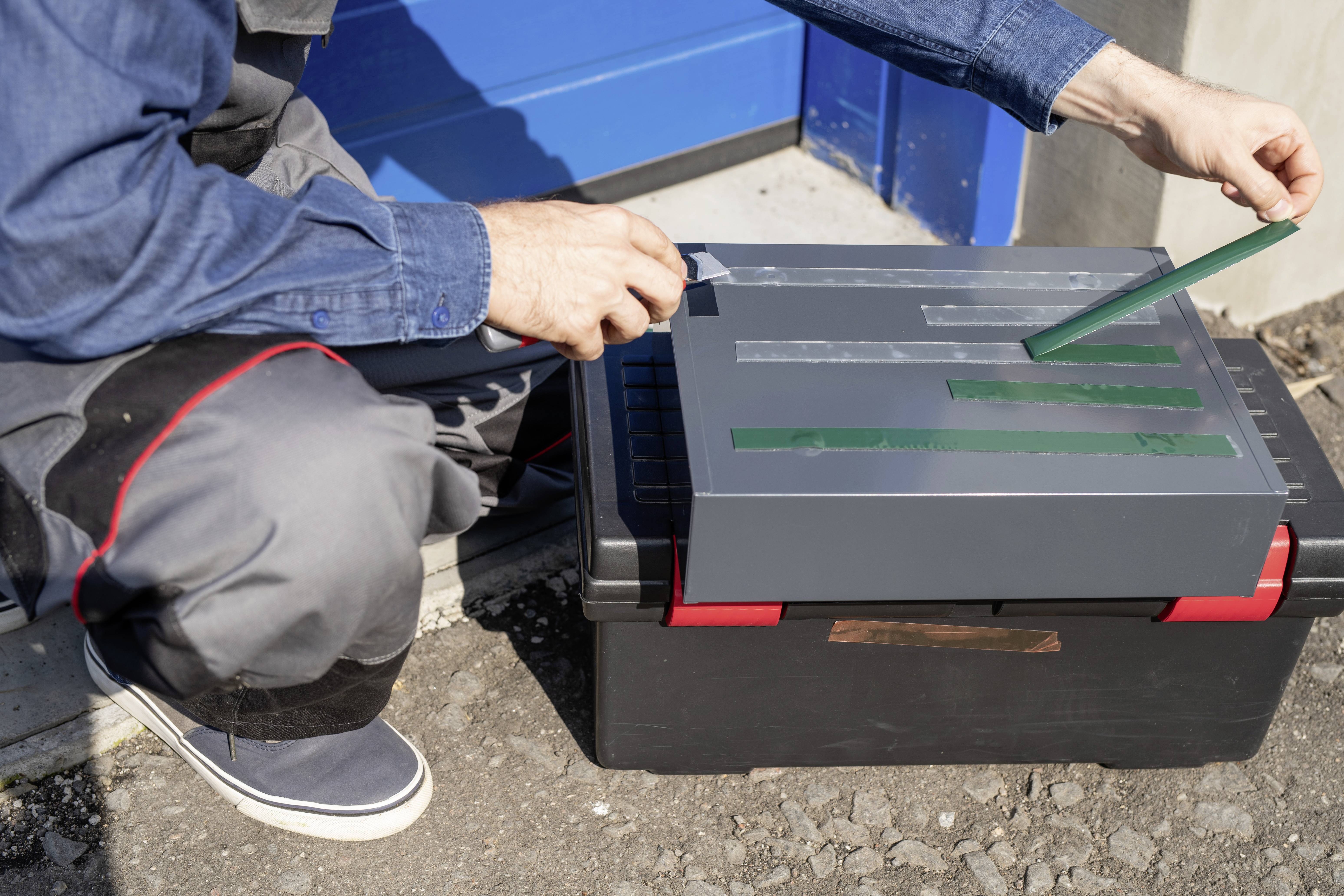 A person is sticking green stripes onto a grey box, which is sitting on a black crate. The background shows a blue door.