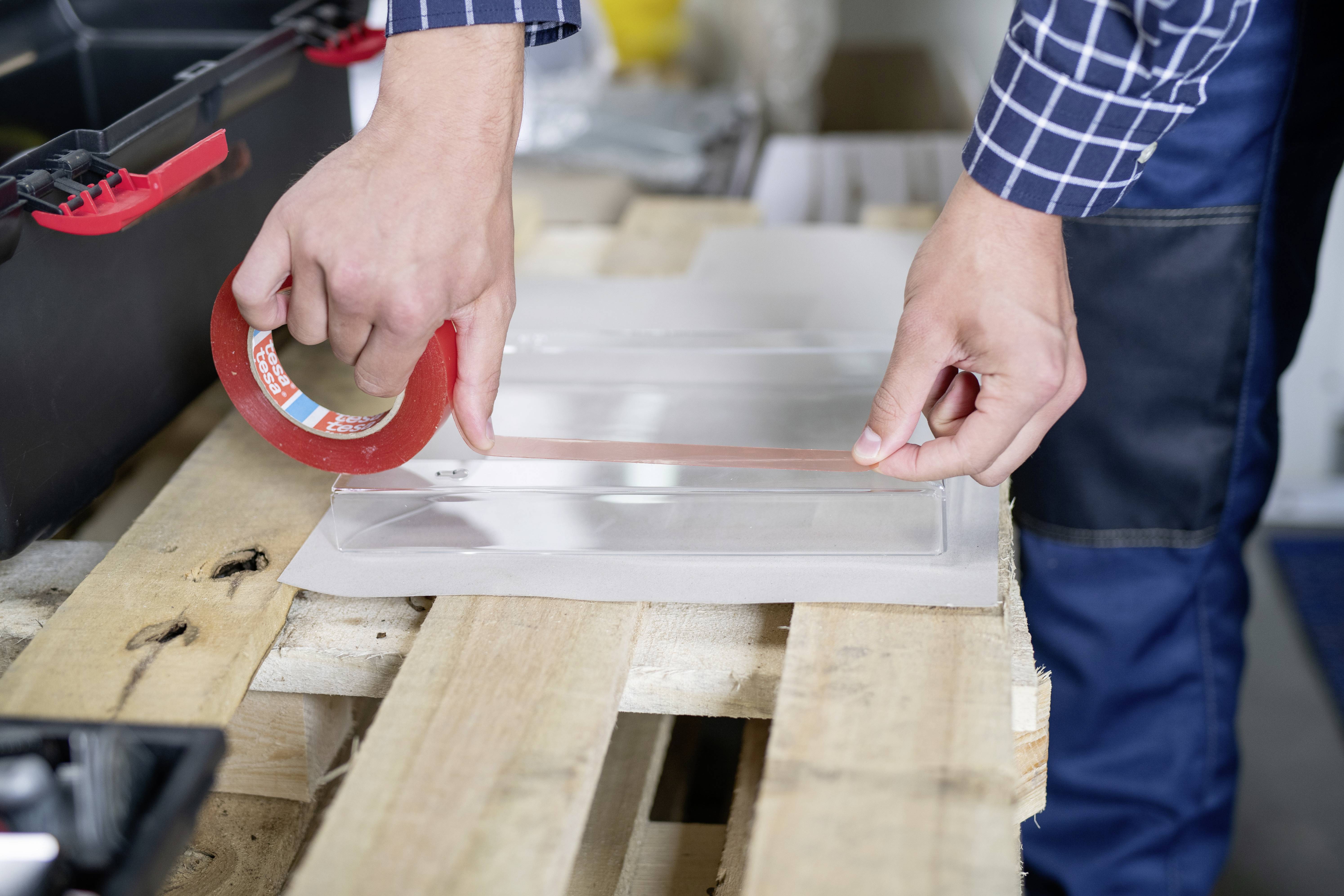 A person is sticking red adhesive tape onto a piece of glass or acrylic lying on a wooden floor. Next to the person is an open toolbox.