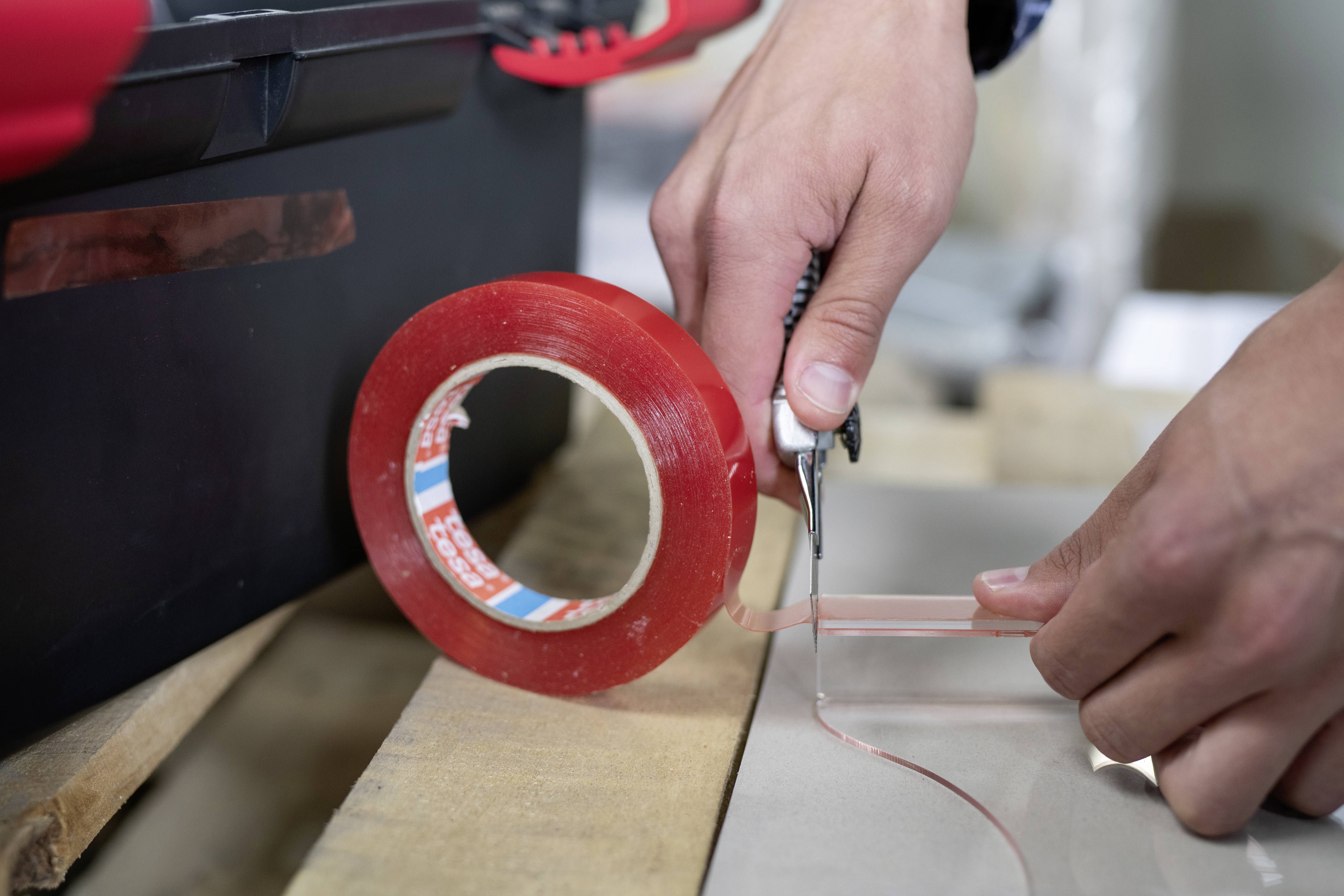 A person cuts a piece of transparent, red adhesive tape from a roll using a Stanley knife on a wooden table.