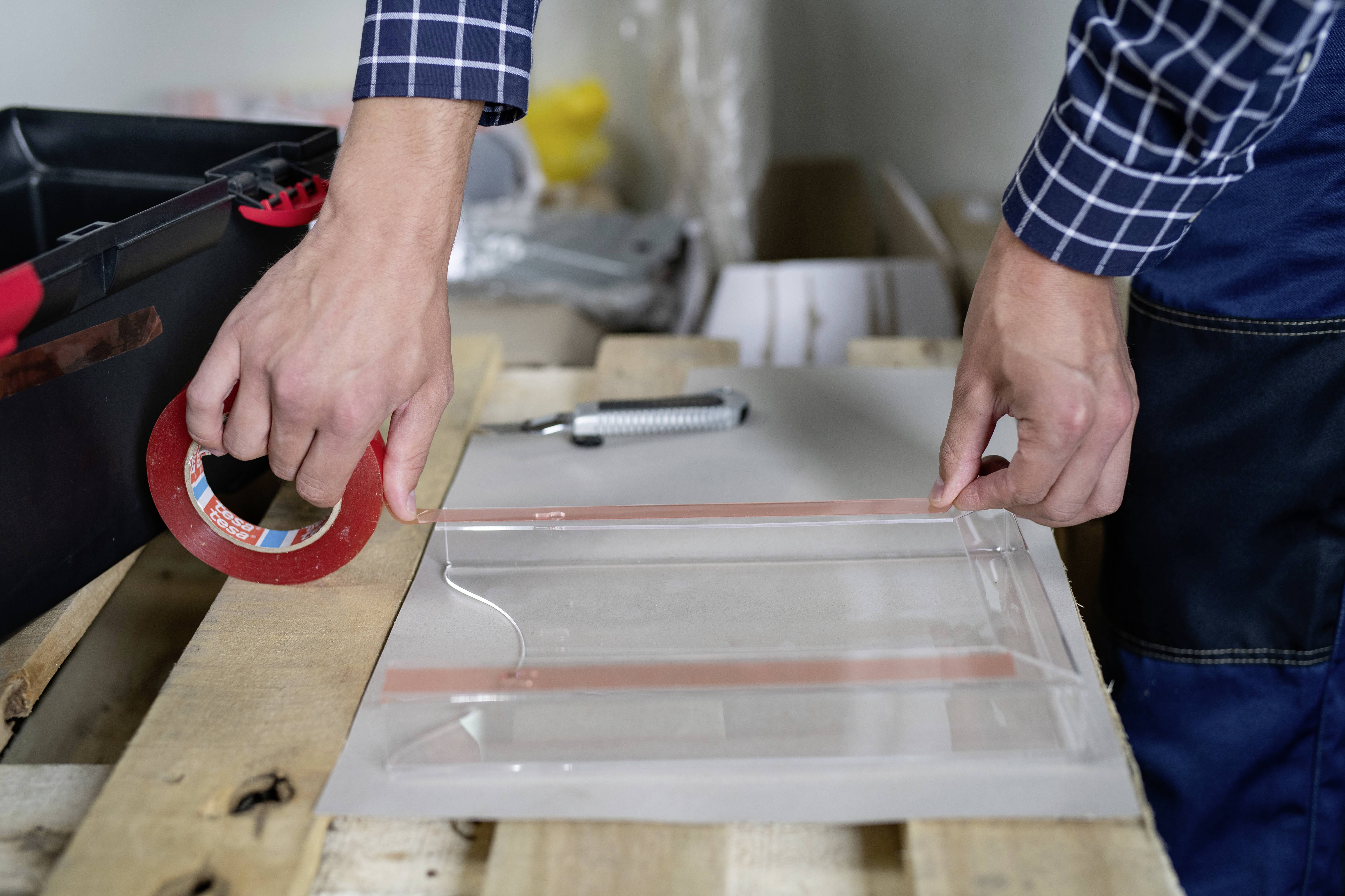 A person is securing a transparent film to wood using adhesive tape. Tools and materials are visible in the background.