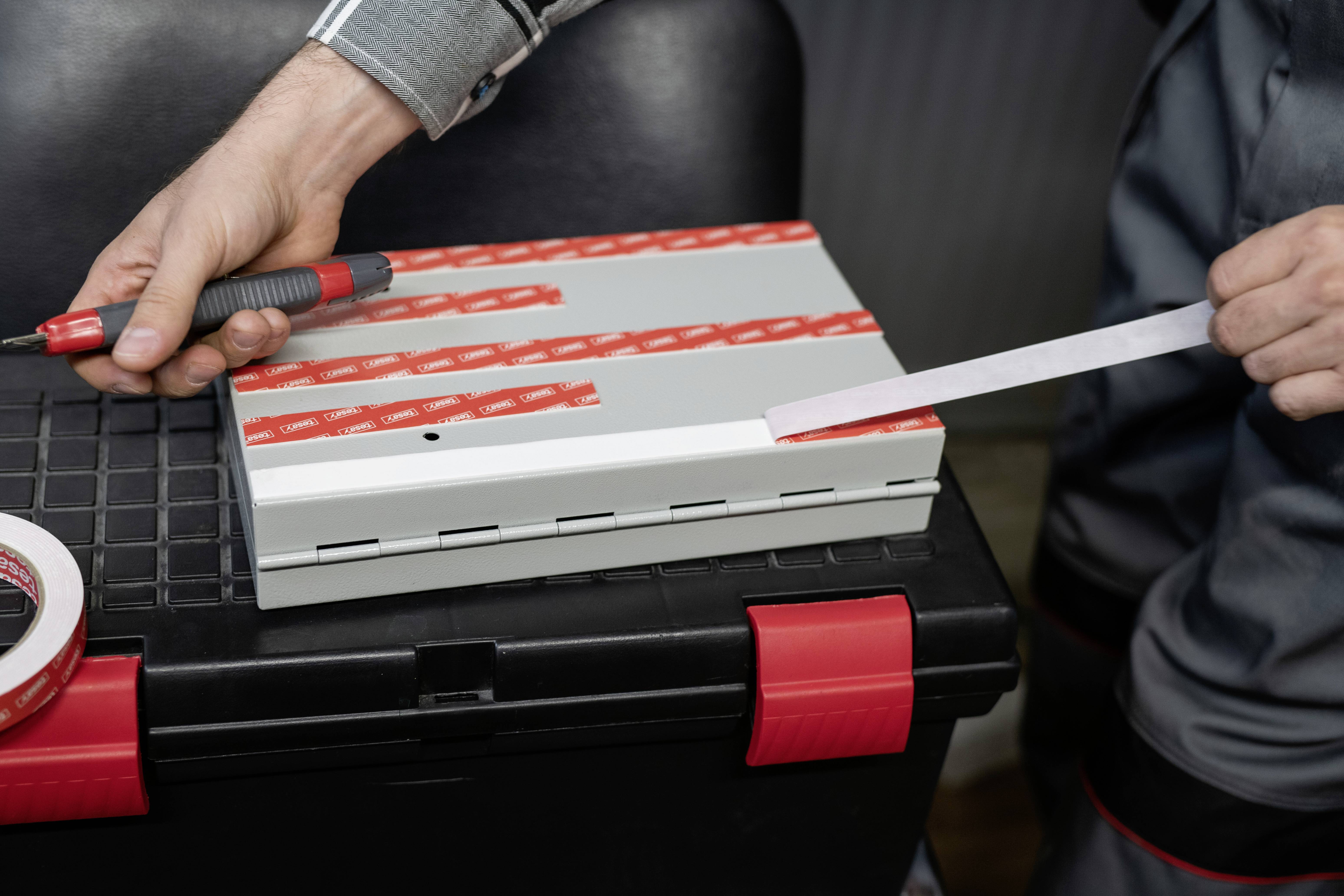 A person is sticking red adhesive strips onto a grey device on a toolbox. They are holding a screwdriver and another roll of tape.