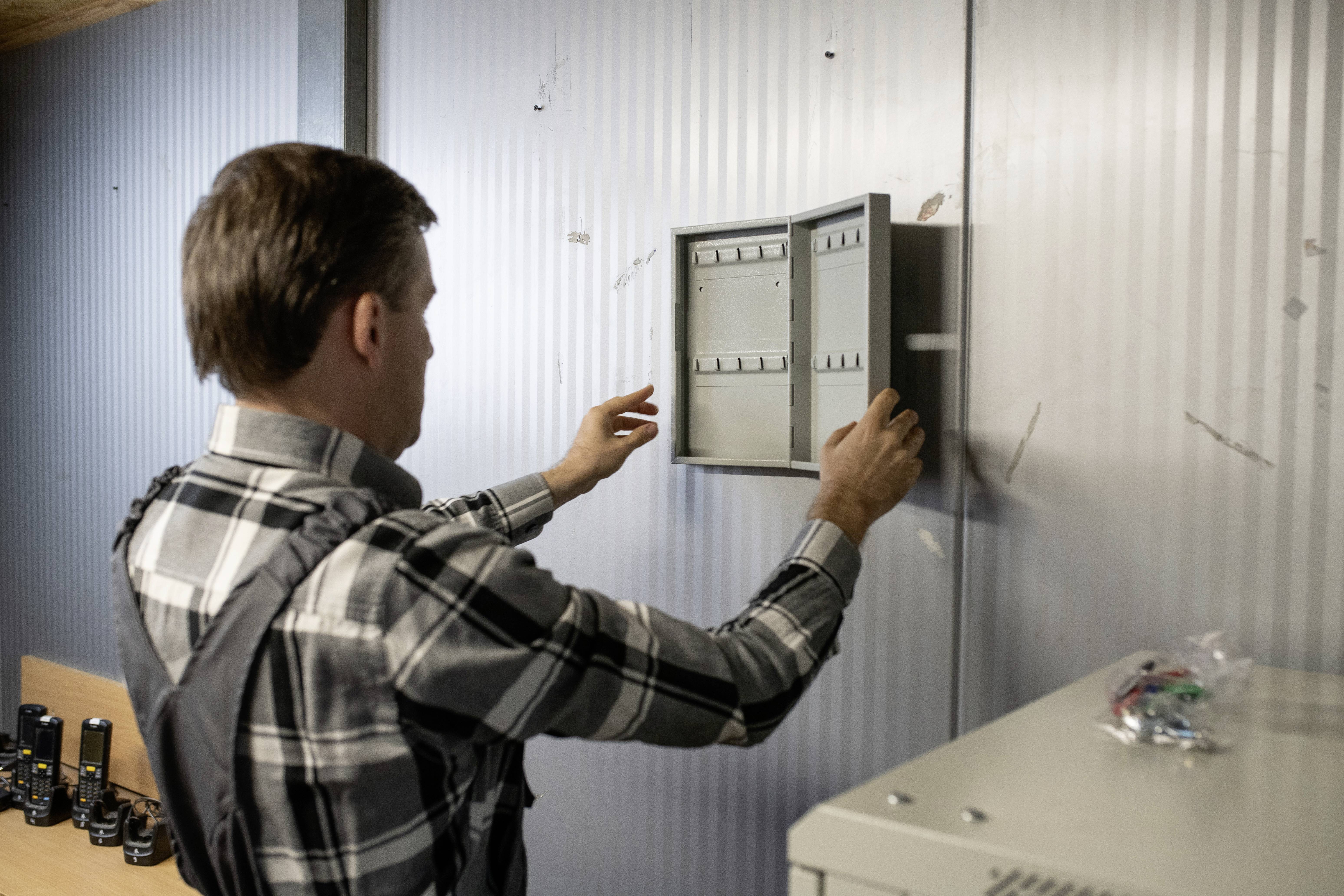 A man opens an empty distribution box on a metal wall in a workshop environment. Tools are lying on a nearby table.