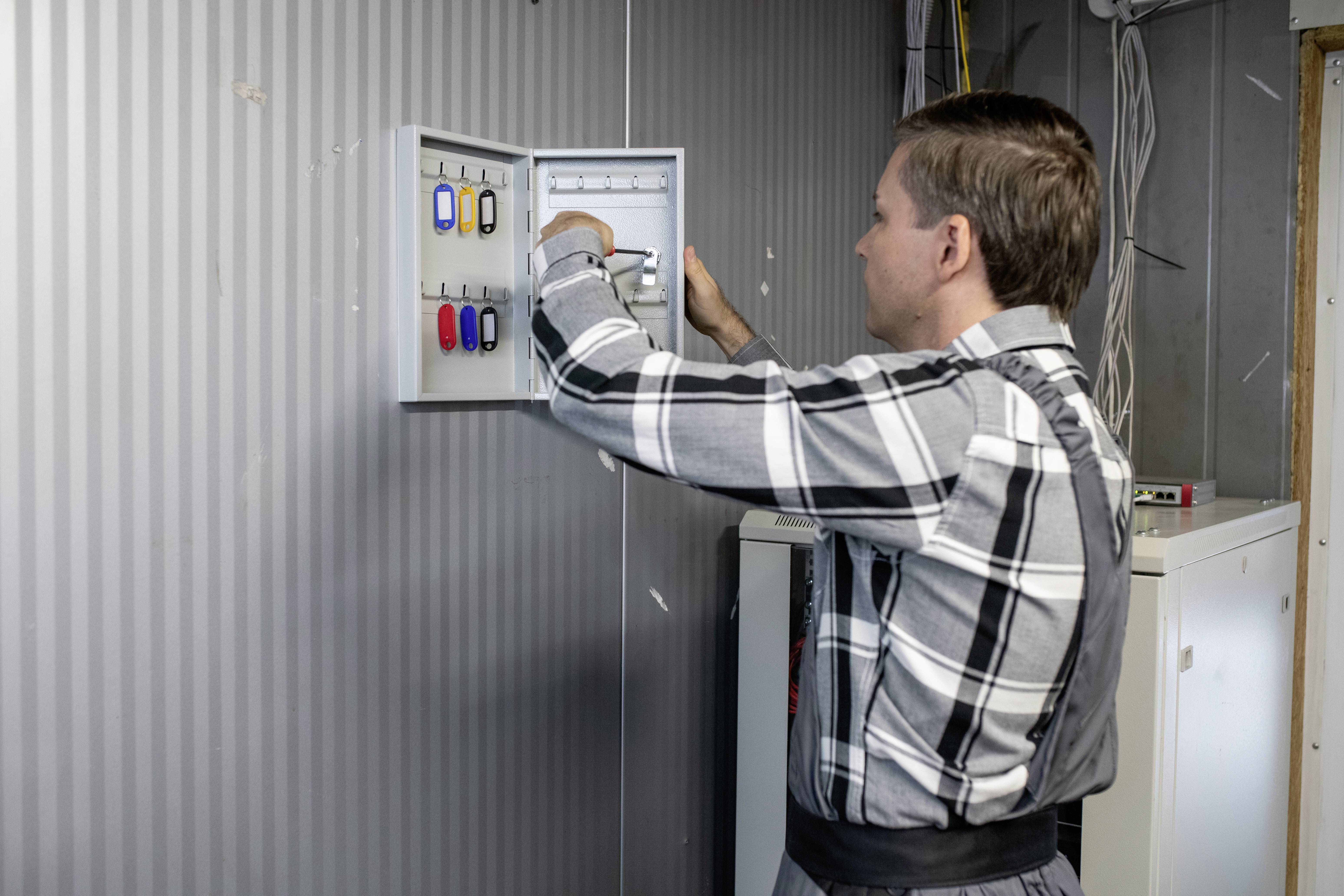 A man in a checked shirt opens a metal cabinet on a wall, which contains various keys with coloured tags.
