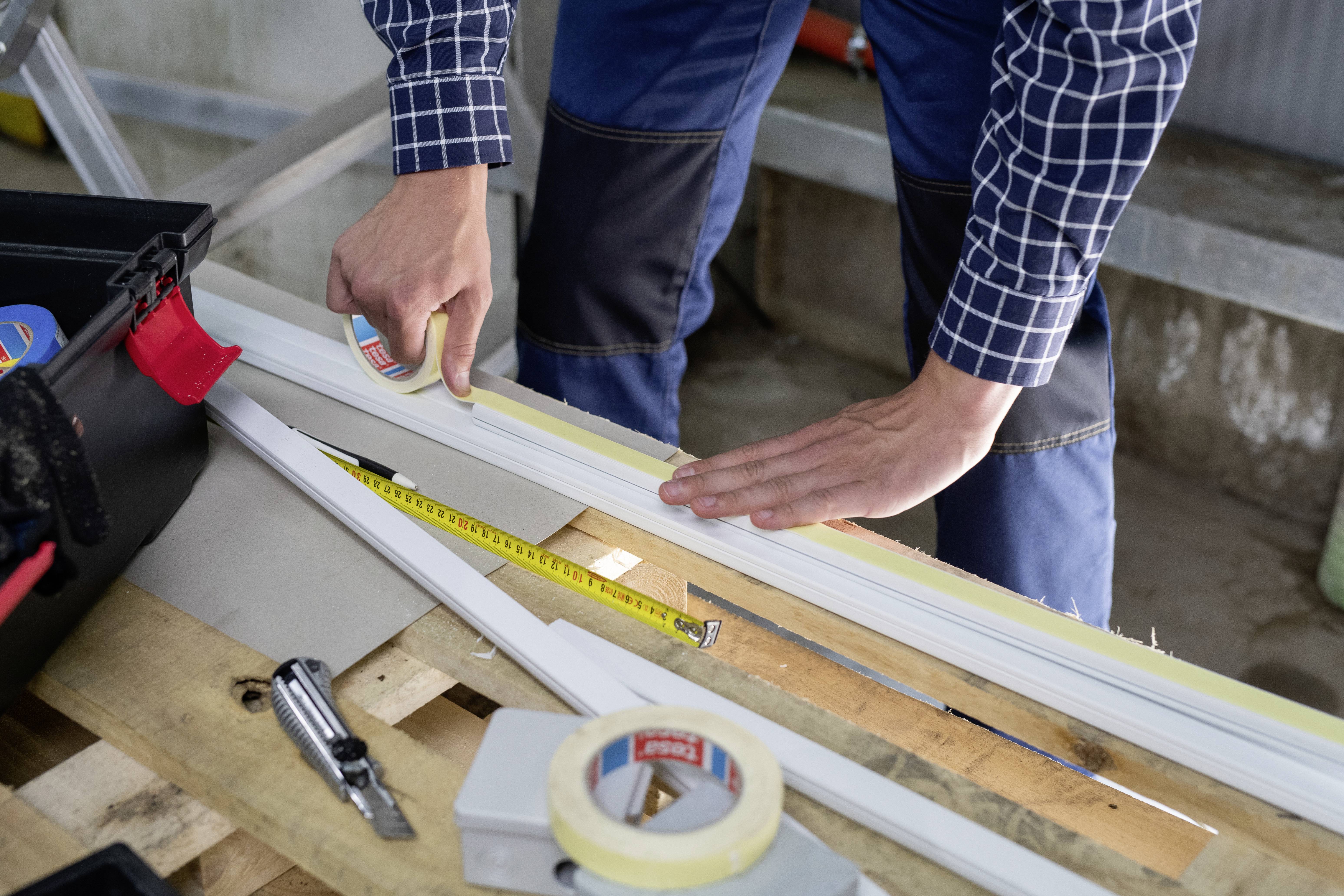 A person is measuring with a tape measure and applying protective tape to a wooden batten on a workbench in a workshop.