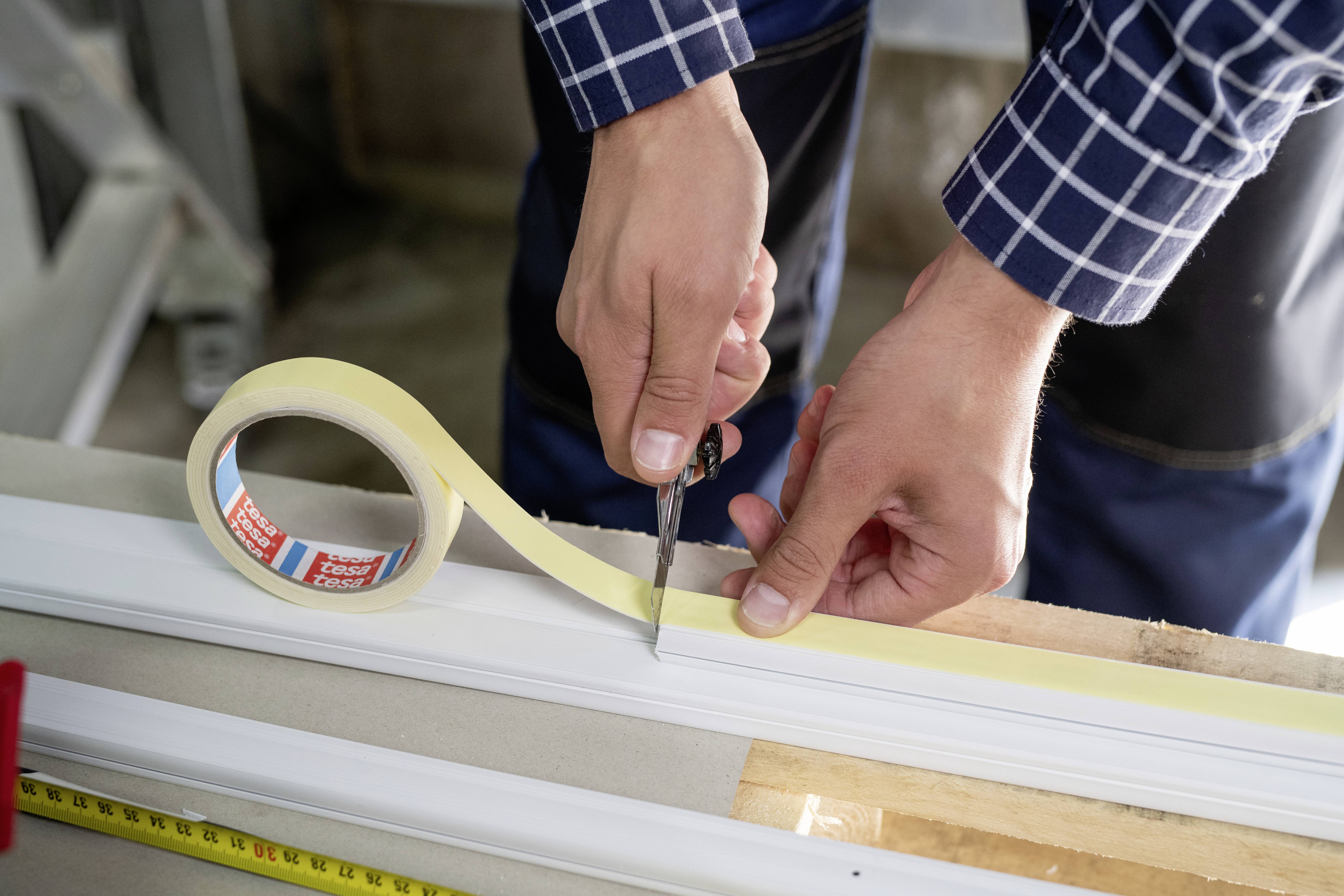 A person is cutting adhesive tape with scissors on a white strip. Wooden tools and a tape measure are visible in the background.