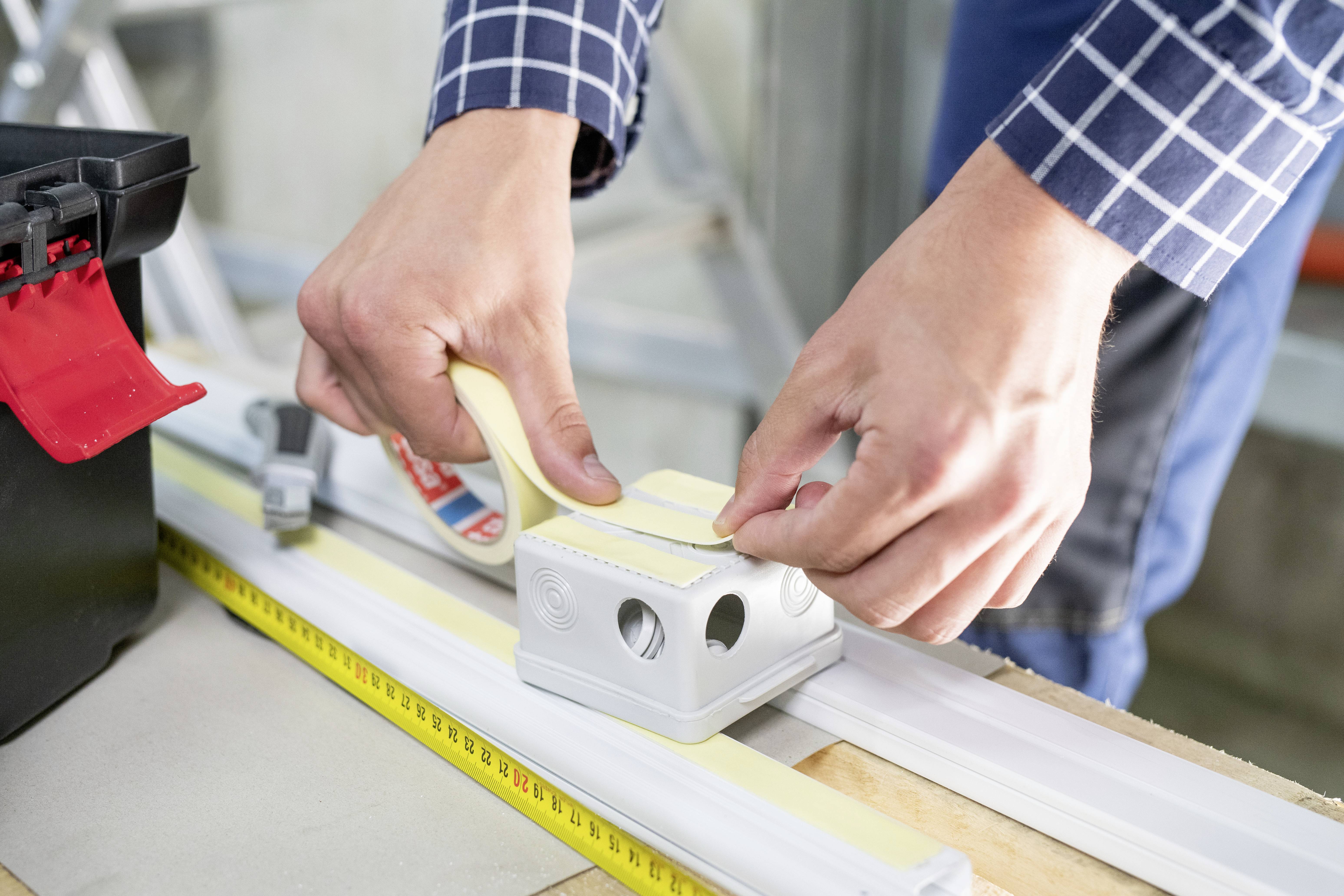 A person is attaching a yellow measuring tape to a white component using double-sided adhesive tape. A toolbox is positioned nearby.
