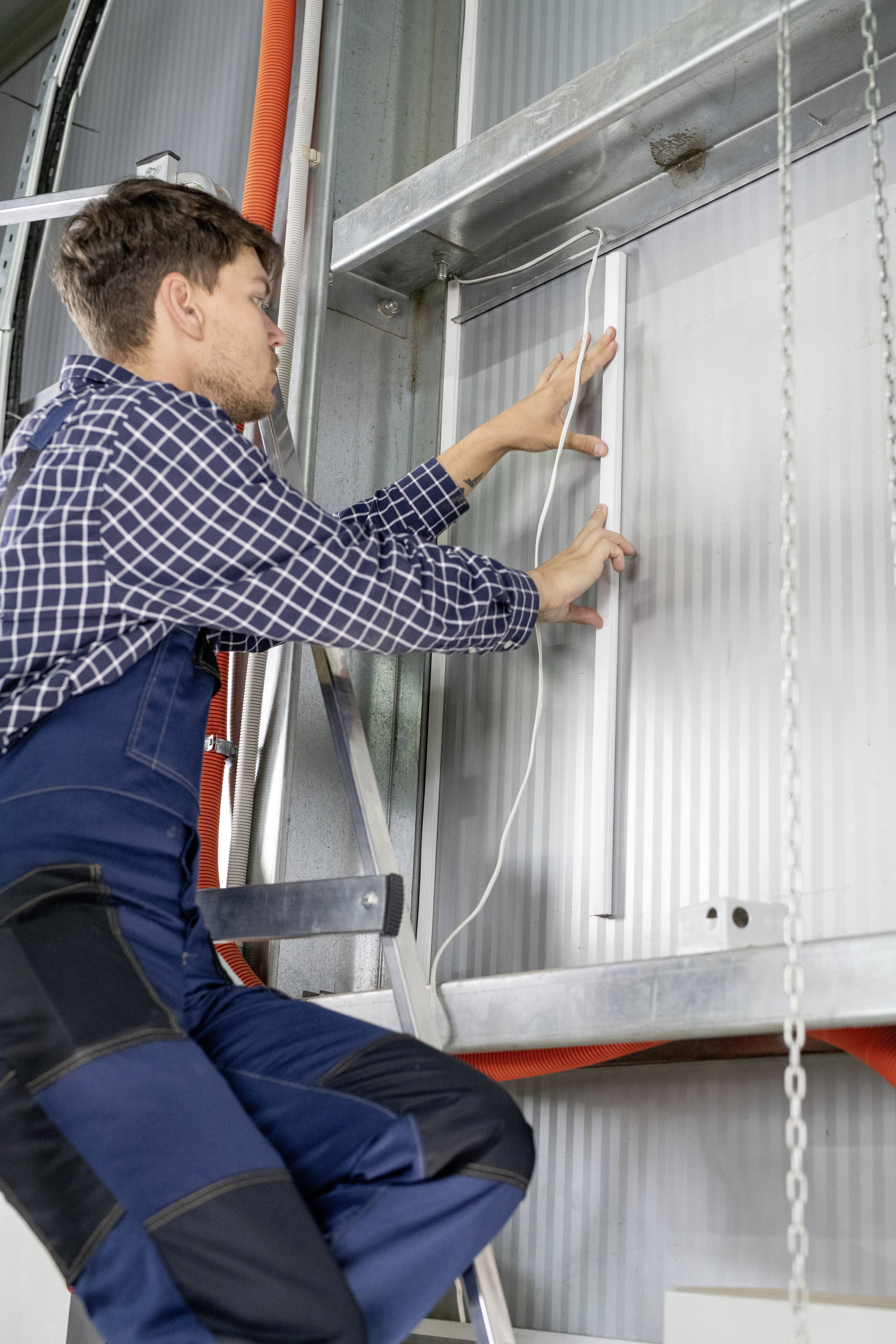 A man in work attire stands on a ladder and measures a metal wall with a tape measure.