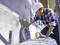 A construction worker wearing a hard hat is attaching cables to a house wall while holding adhesive tape in his hand.