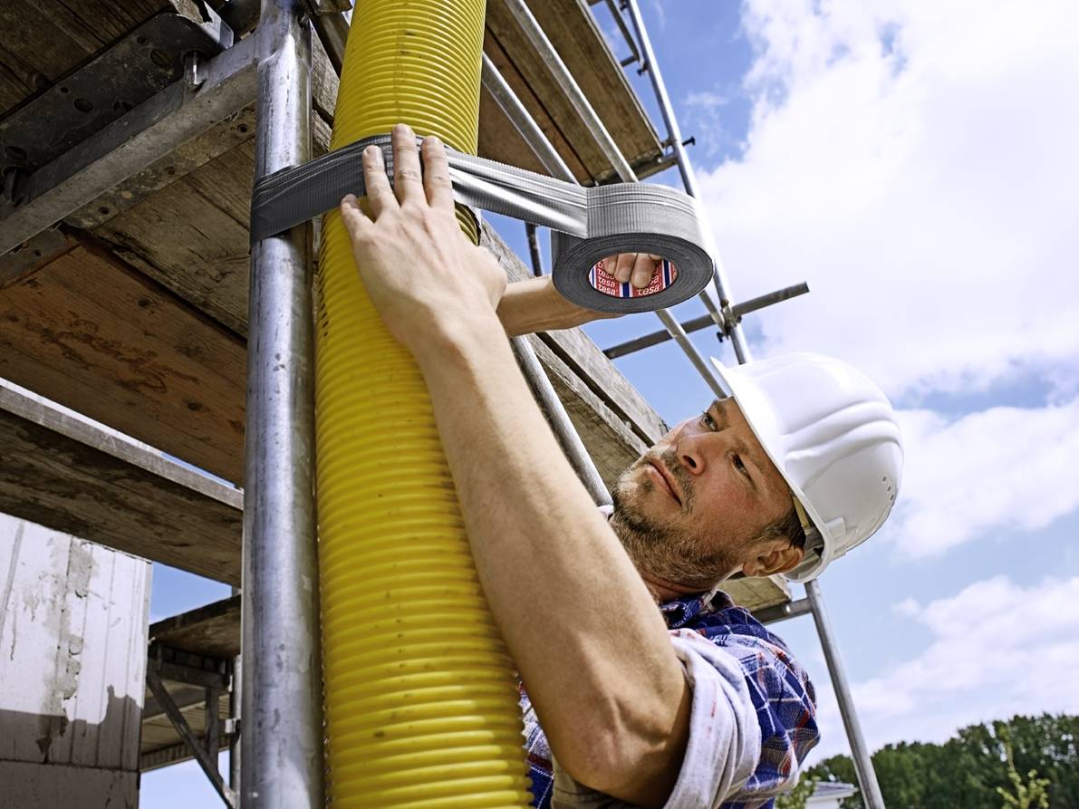 A construction worker wearing a hard hat is securing a yellow pipe to scaffolding with tape. Parts of the blue sky are visible in the background.