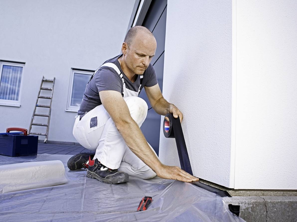 A tradesman is applying a black sealing strip to the underside of a house wall. A ladder is leaning against the wall in the background.