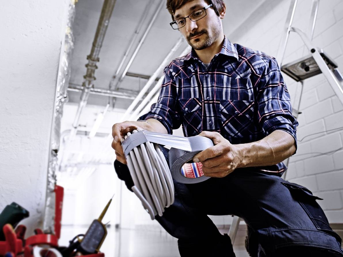 A man in workwear is inspecting a cable bundle in an industrial setting. A ladder is visible in the background.