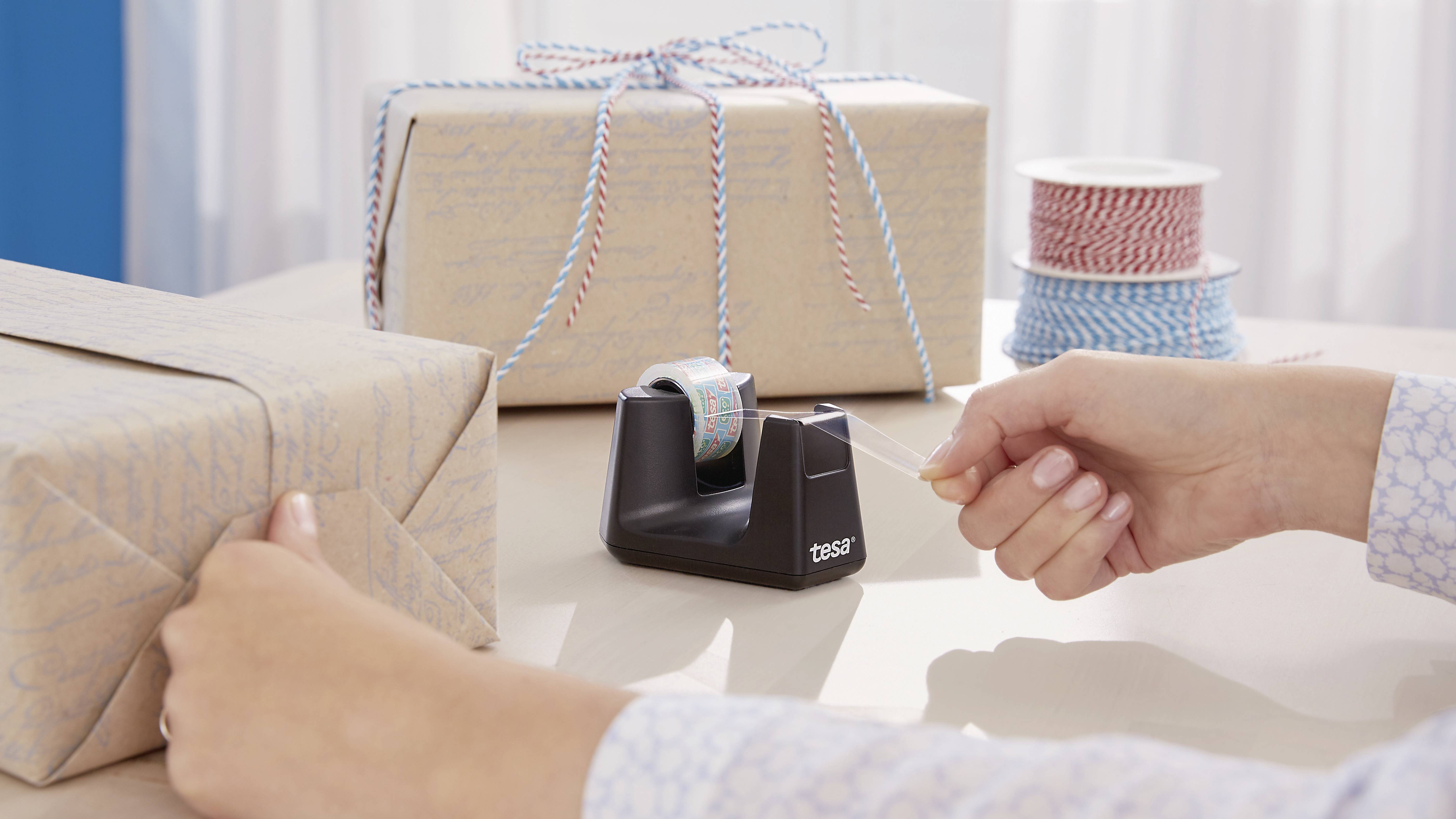 Two wrapped gifts with ribbons. One person is cutting tape from a black tape dispenser. Background: colourful strings.