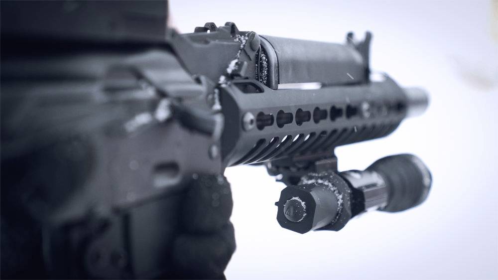 A close-up of a blackened rifle barrel with a telescopic sight, partially covered in snow.