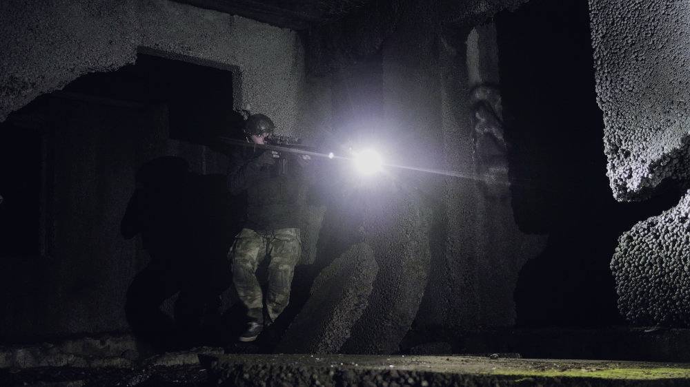 A person in camouflage stands in a dark, dilapidated building and shines a torch on something in the foreground.
