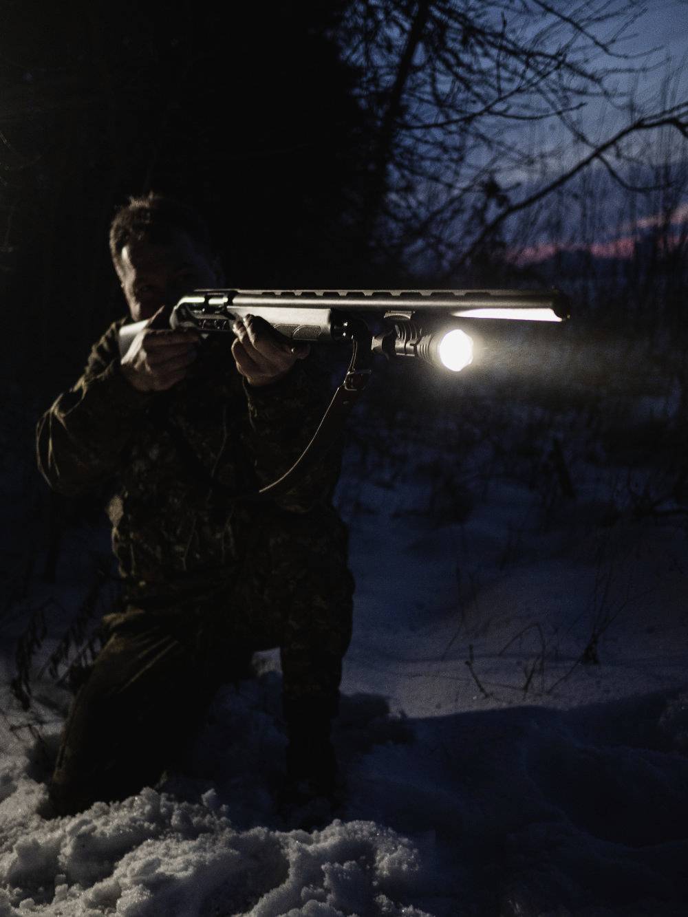 A person is kneeling in the snow and aiming a rifle with an attached torch in a dark, wooded environment.