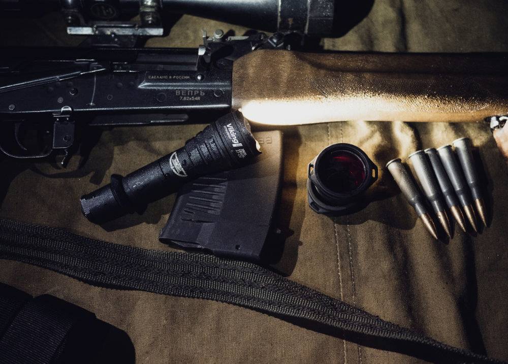 A weapon with a wooden stock lies on a table, surrounded by a torch, a magazine, a lens, and five rounds.