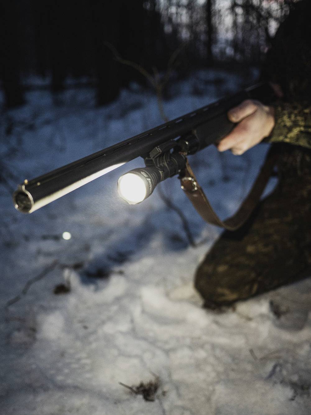 A person is kneeling in the snow, holding a rifle with a torch. The surroundings appear dark and wintry, presumably in a forest.
