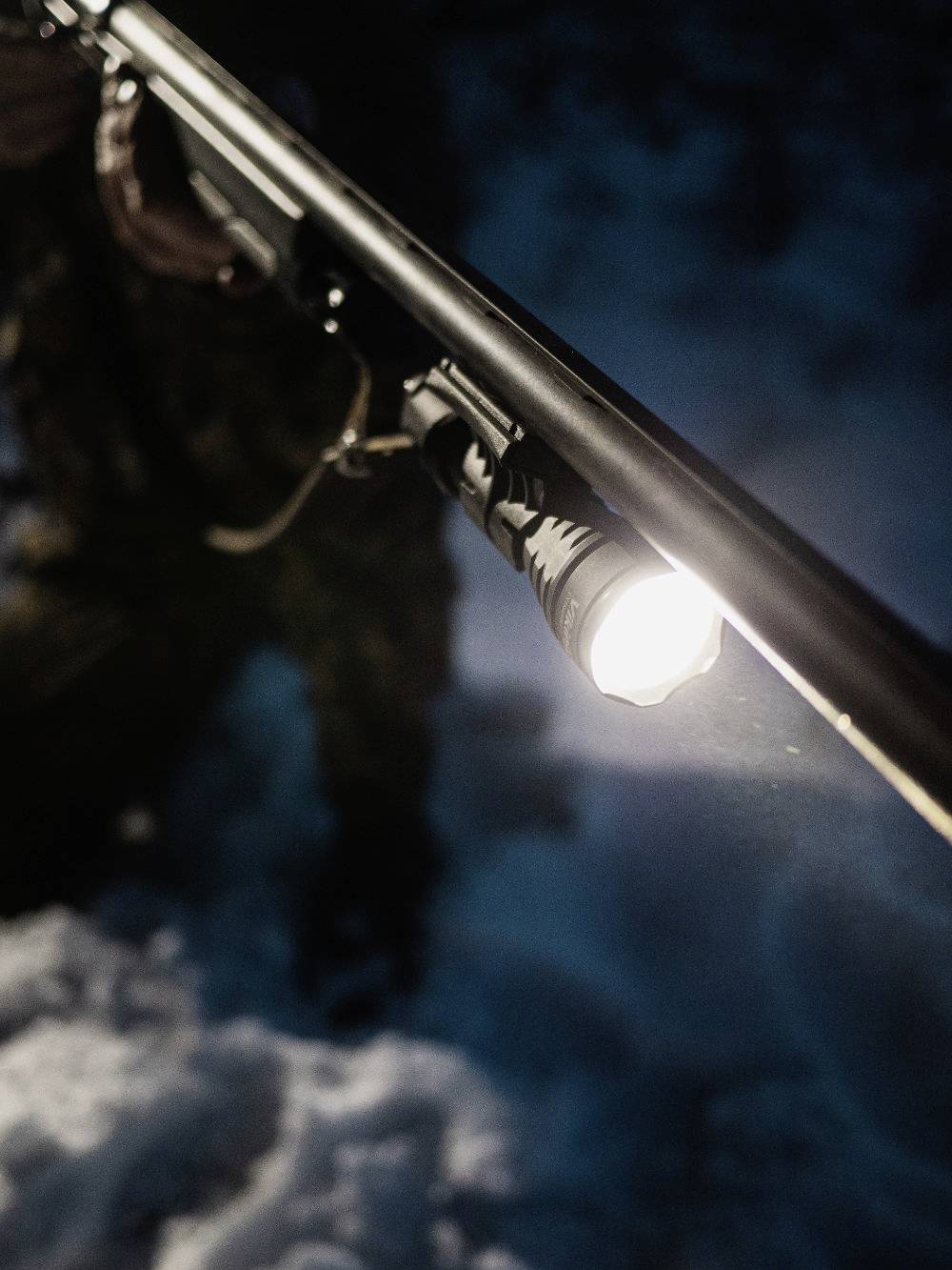 A bright torch attached to a gun barrel in the darkness illuminates a snow-covered ground.