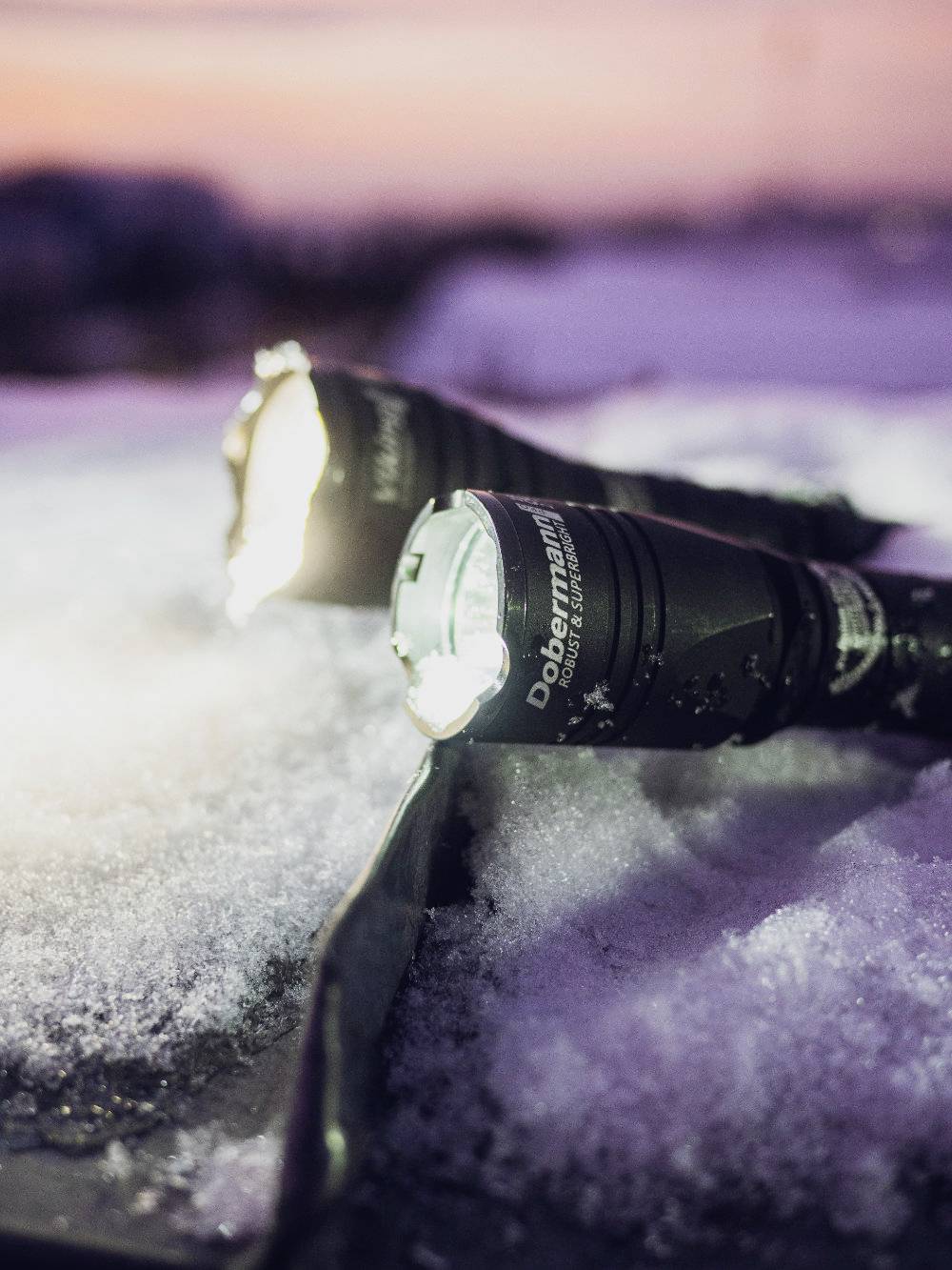 Two torches shine in the snow at sunset. The front one is switched on and beams brightly onto the crystalline surface.