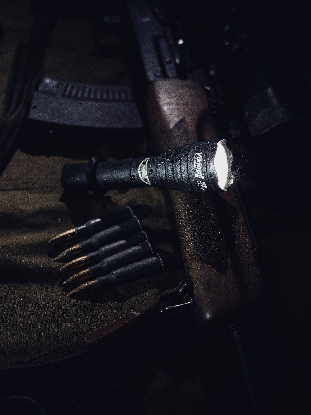 Torch illuminates a rifle and cartridges lying on a cloth. The scene is dark, focused on the weapons.