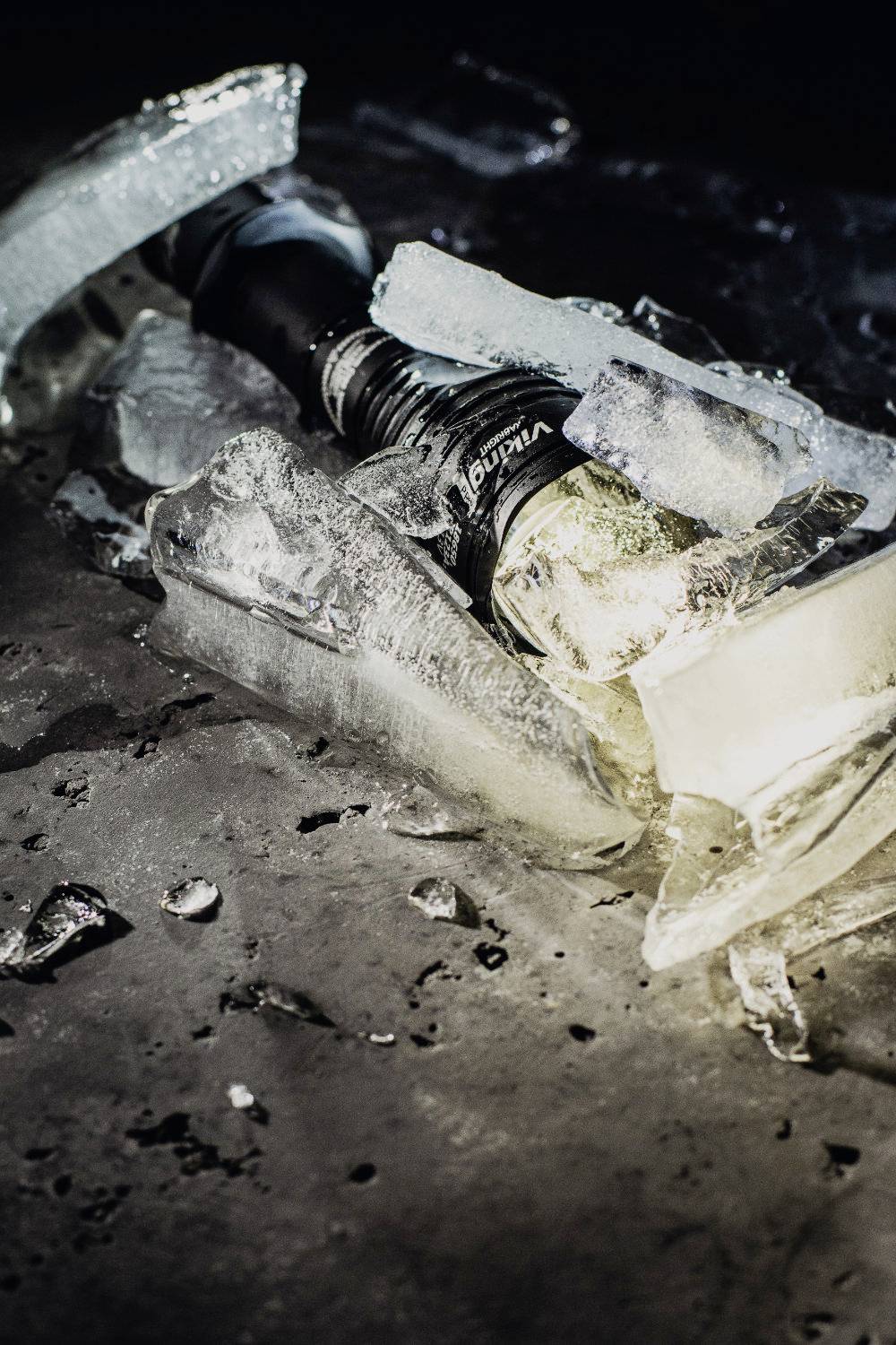 A broken light bulb lies on a dark, ice-covered surface. The pieces of ice are unevenly distributed around the bulb.