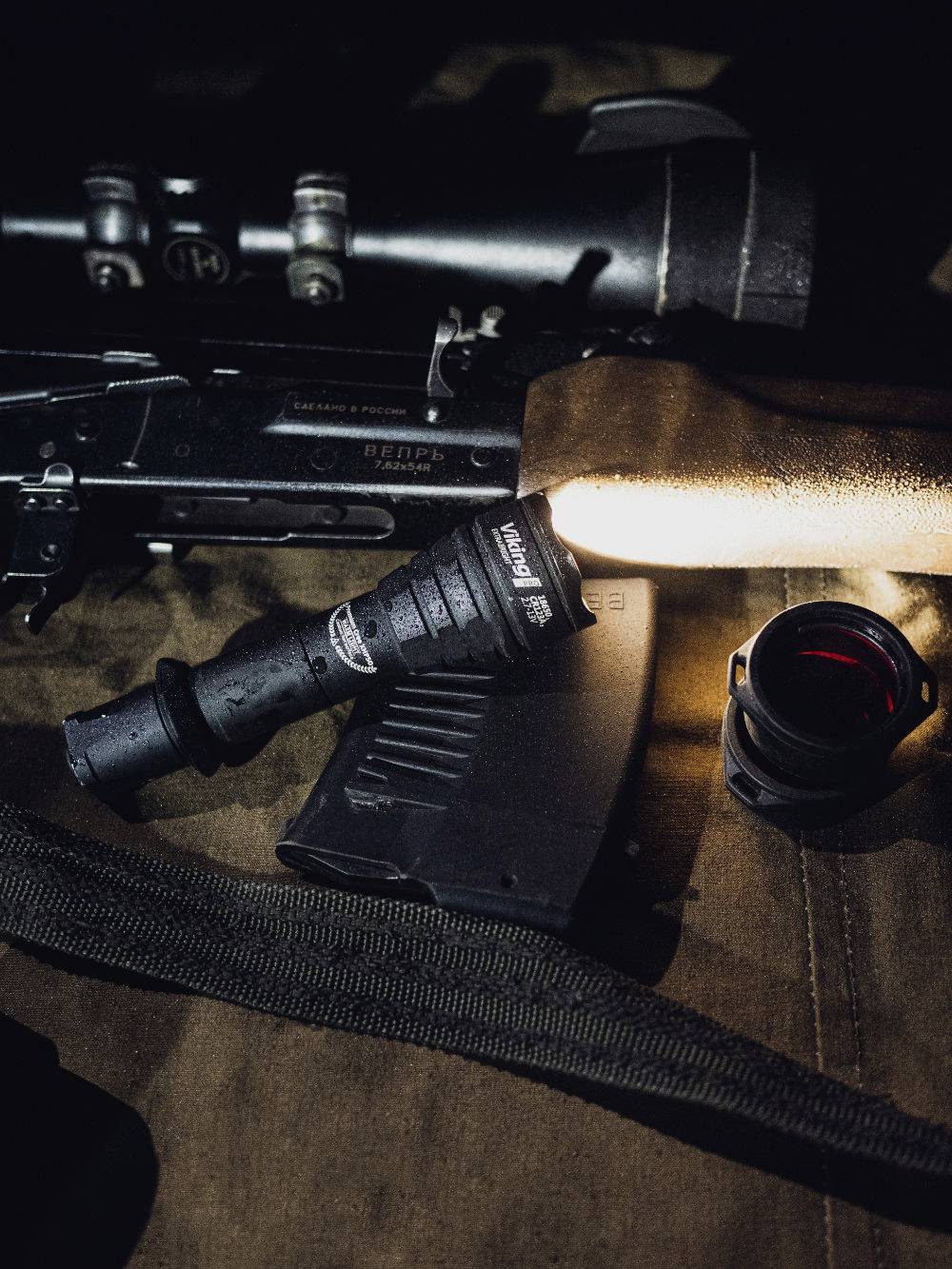 A tactical torch illuminates a rifle magazine, alongside a telescopic sight and a lens, all on a brown surface.