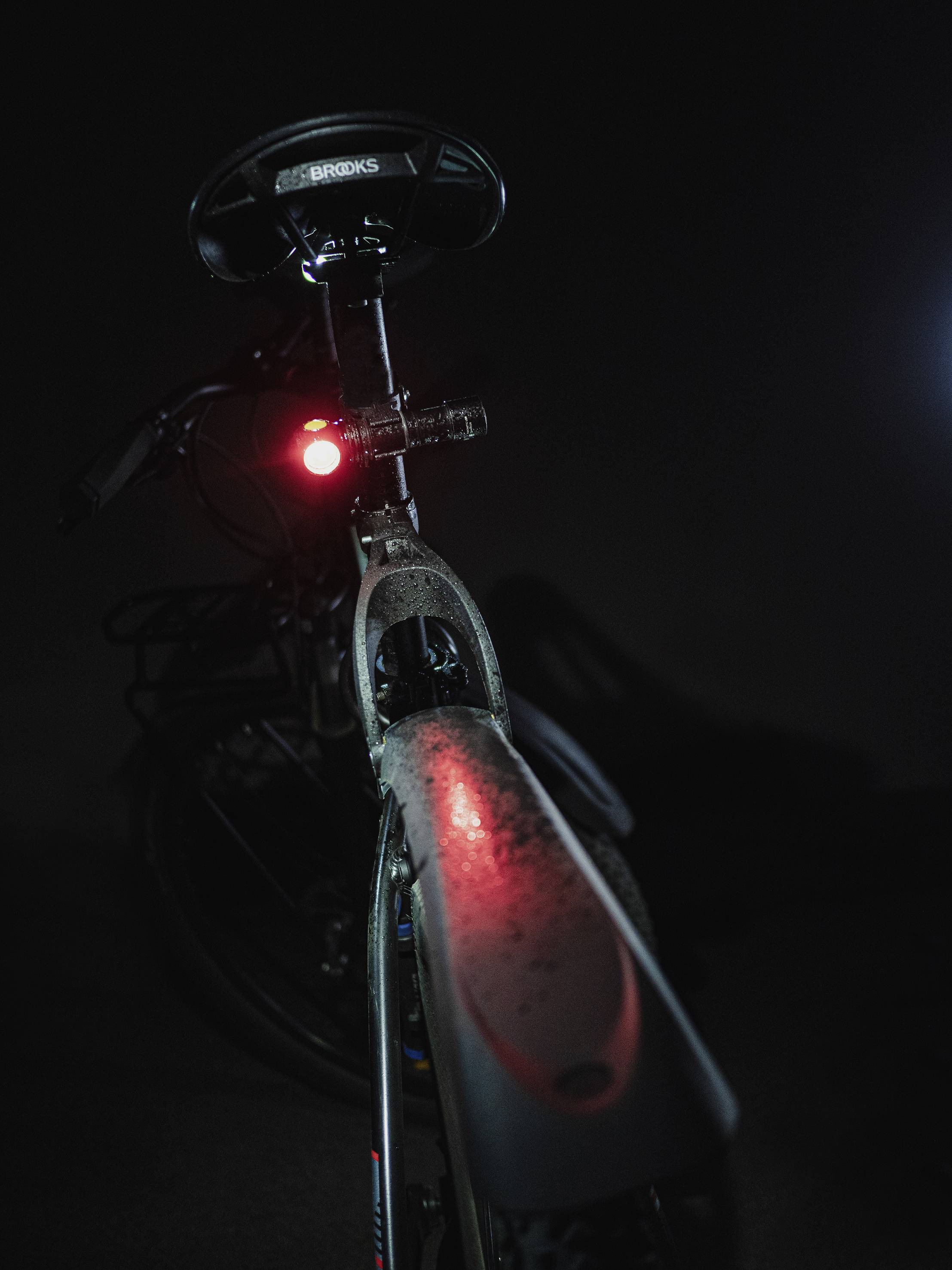 A bicycle from behind in dim light, with the saddle and red rear light visible. The background is dark.