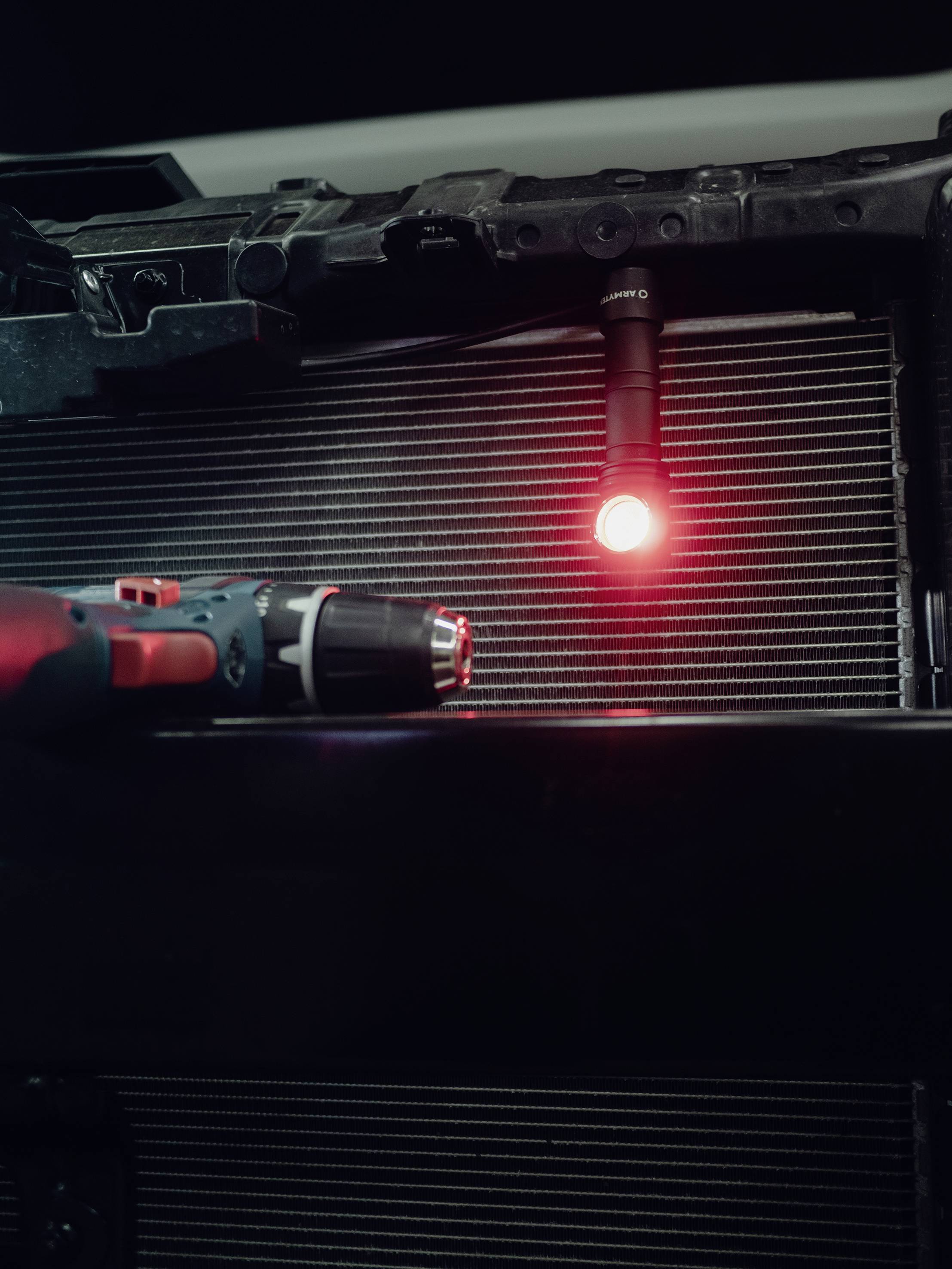 A red LED light illuminates the area in front of a metallic radiator grille. In the foreground, a cordless drill moves.