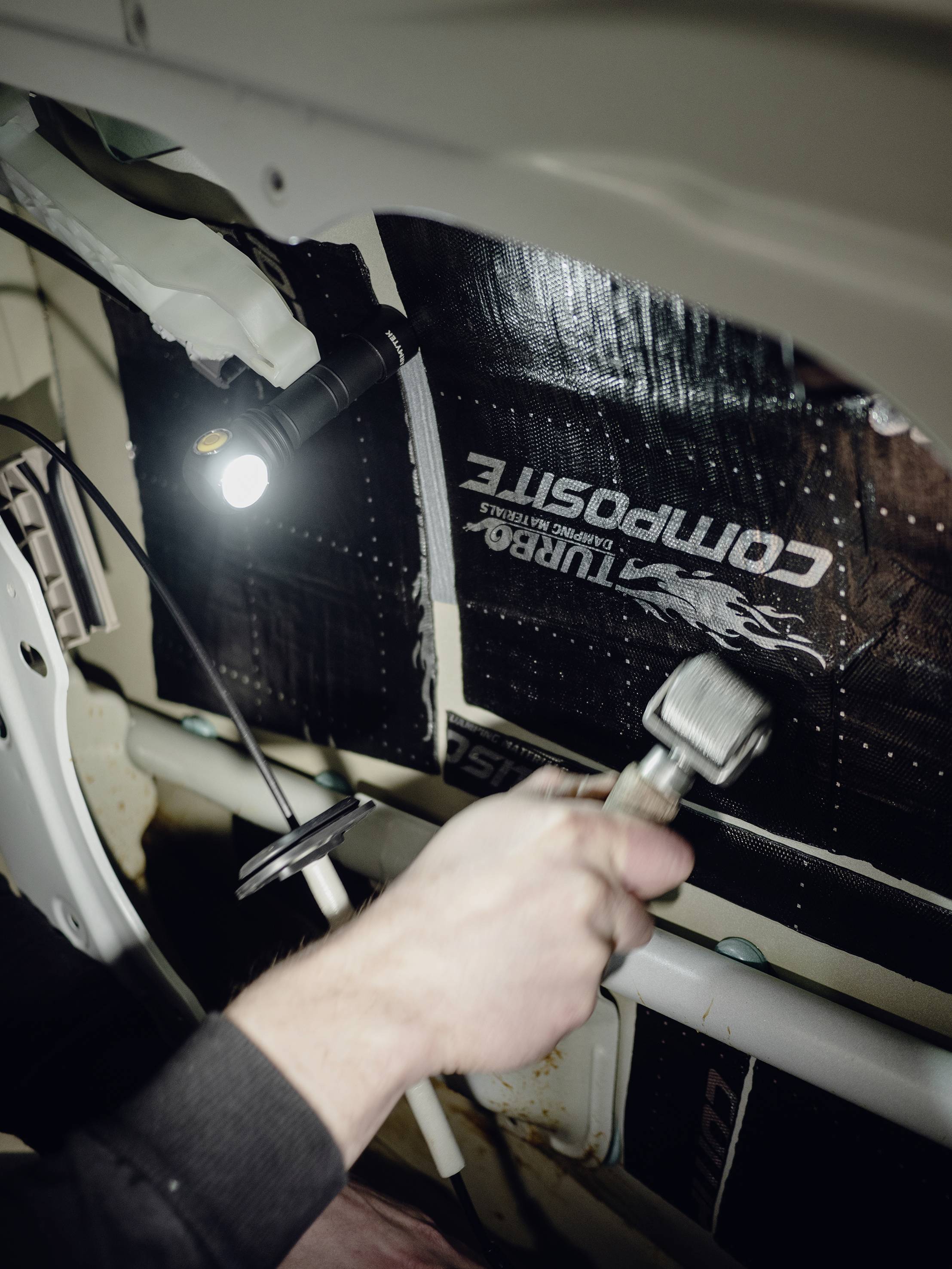 A person in a vehicle is working with a tool on the interior insulation, illuminated by a lamp.